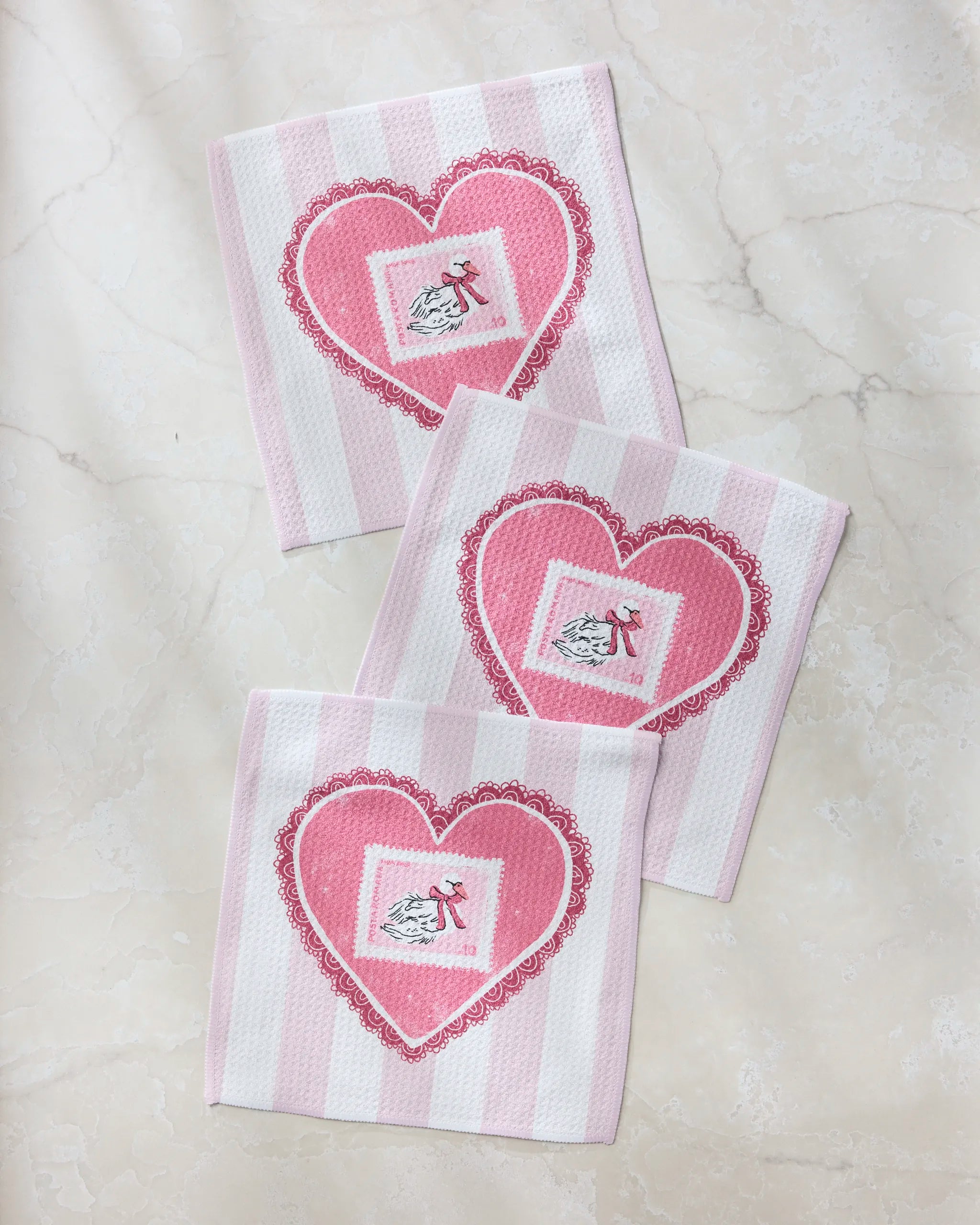 The Geometry Love and Swans dishcloth set includes three eco-friendly, pink and white striped cloths with heart designs, each accented by a bird and bow—perfect for adding style to any kitchen.