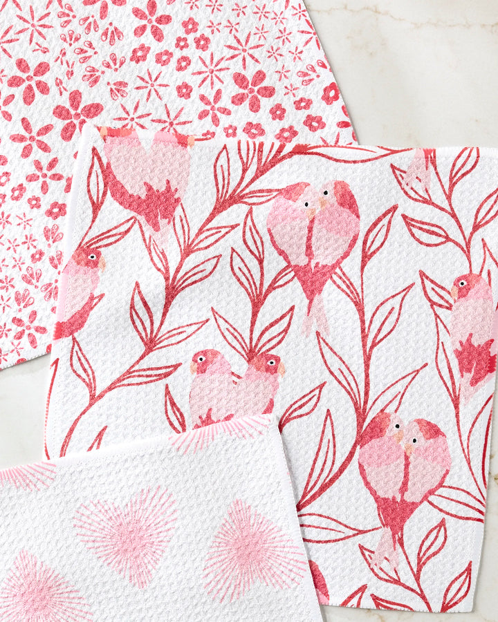 The Love Birds dishcloth set from Hufton Studio includes three super-absorbent towels with pink and red flowers, birds on branches, and pink hearts—perfect for brightening your kitchen.