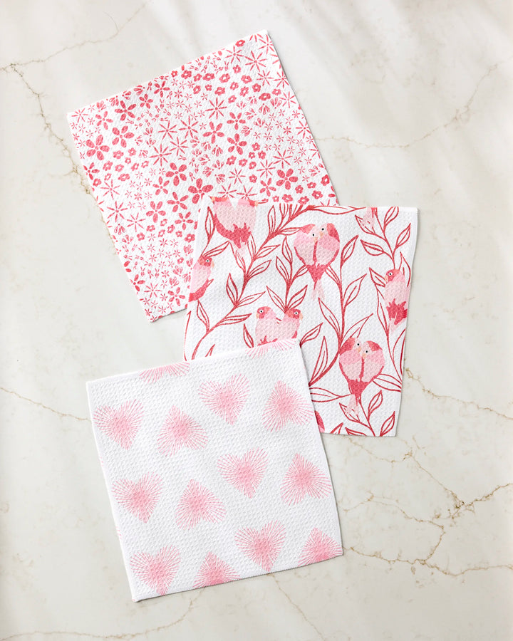 Three "Love Birds" square paper napkins by Hufton Studio, featuring pink floral, bird, and heart patterns, are displayed on marble beside a set of super-absorbent dishtowels made from recycled materials.