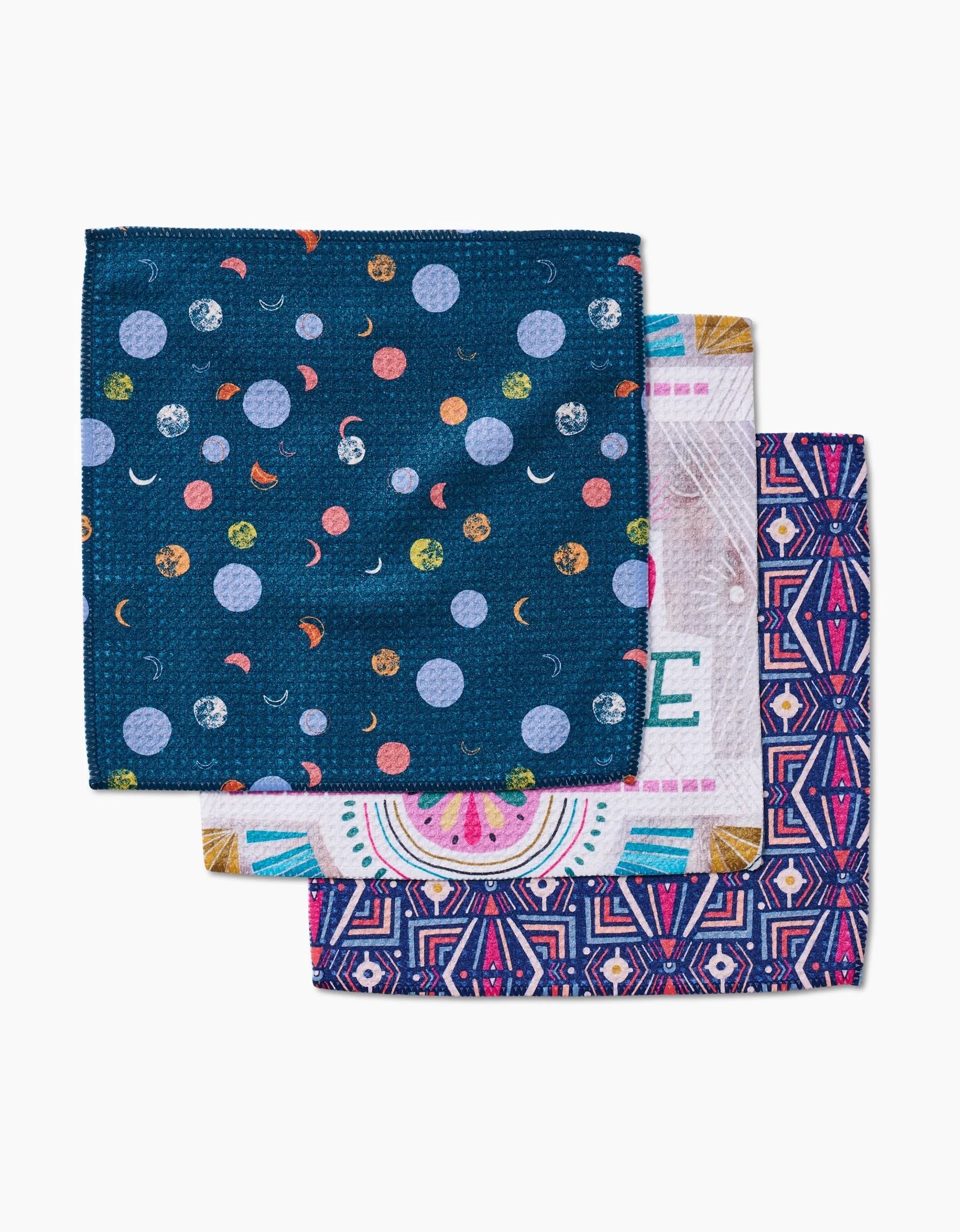 The Jessica Swift Love is Good Medicine set includes three square dishcloths: one with planets on dark blue, one with pastel text designs, and one with a geometric purple, pink, and white pattern—all in Geoweave fabric.