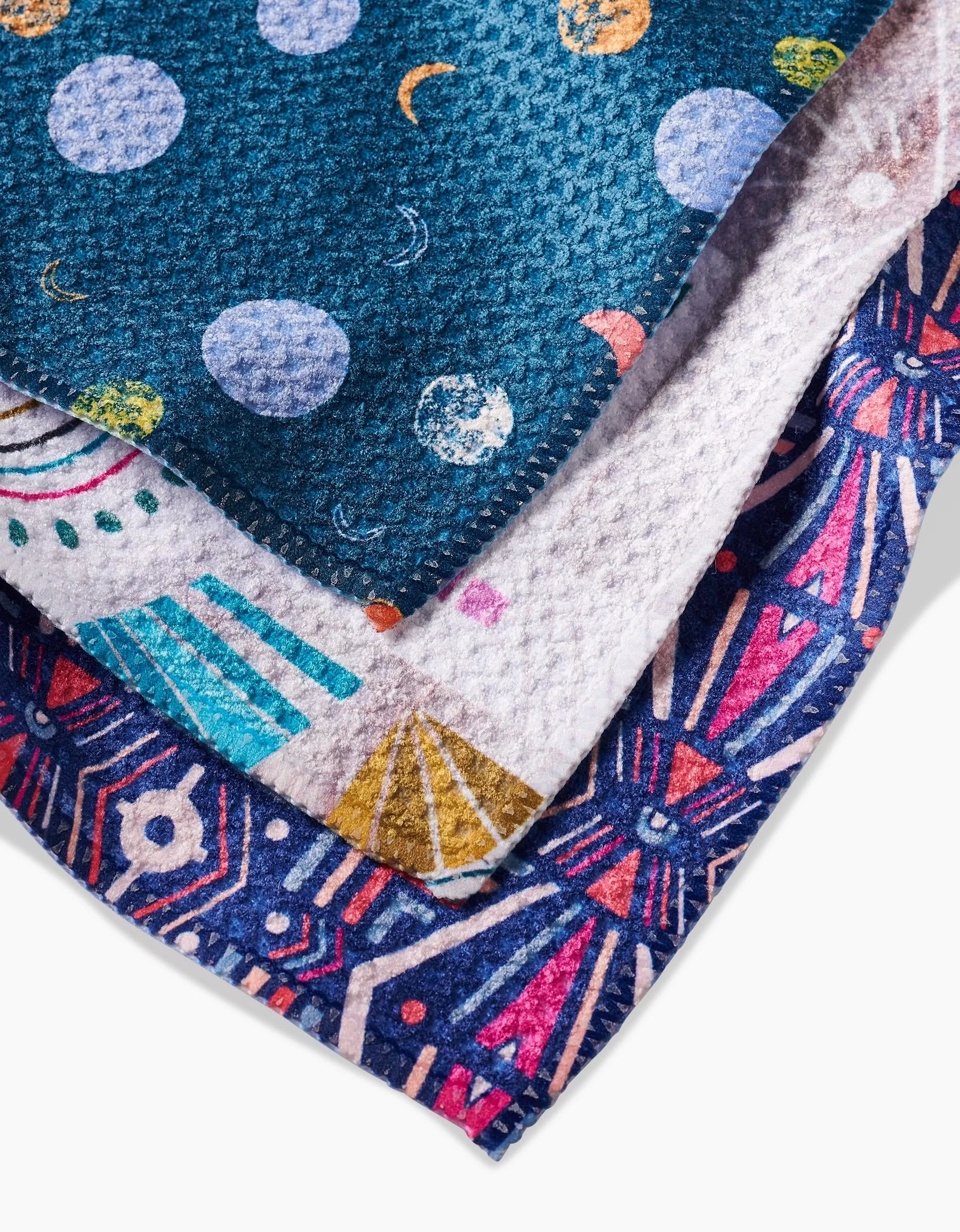 The Love is Good Medicine dishcloth set by Jessica Swift includes three eco-friendly cloths: one with planets and moons, one with colorful shapes, and one with a bold geometric pattern—all made from recycled materials.