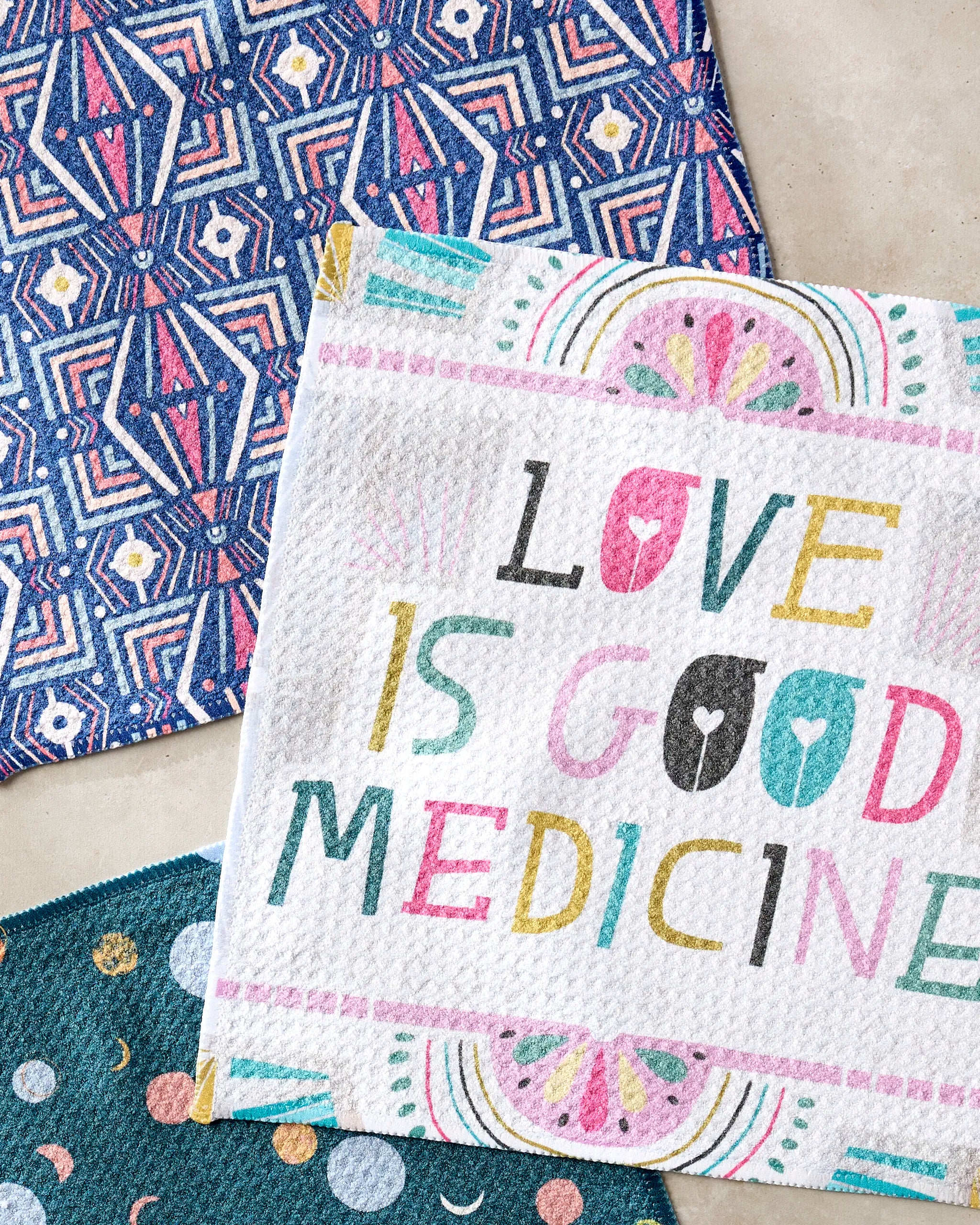 The "Love is Good Medicine" dishcloth set by Jessica Swift includes two super-absorbent towels—one with blue geometric patterns, one with blue circles and moons—and a recycled material cloth featuring the phrase in playful fonts.