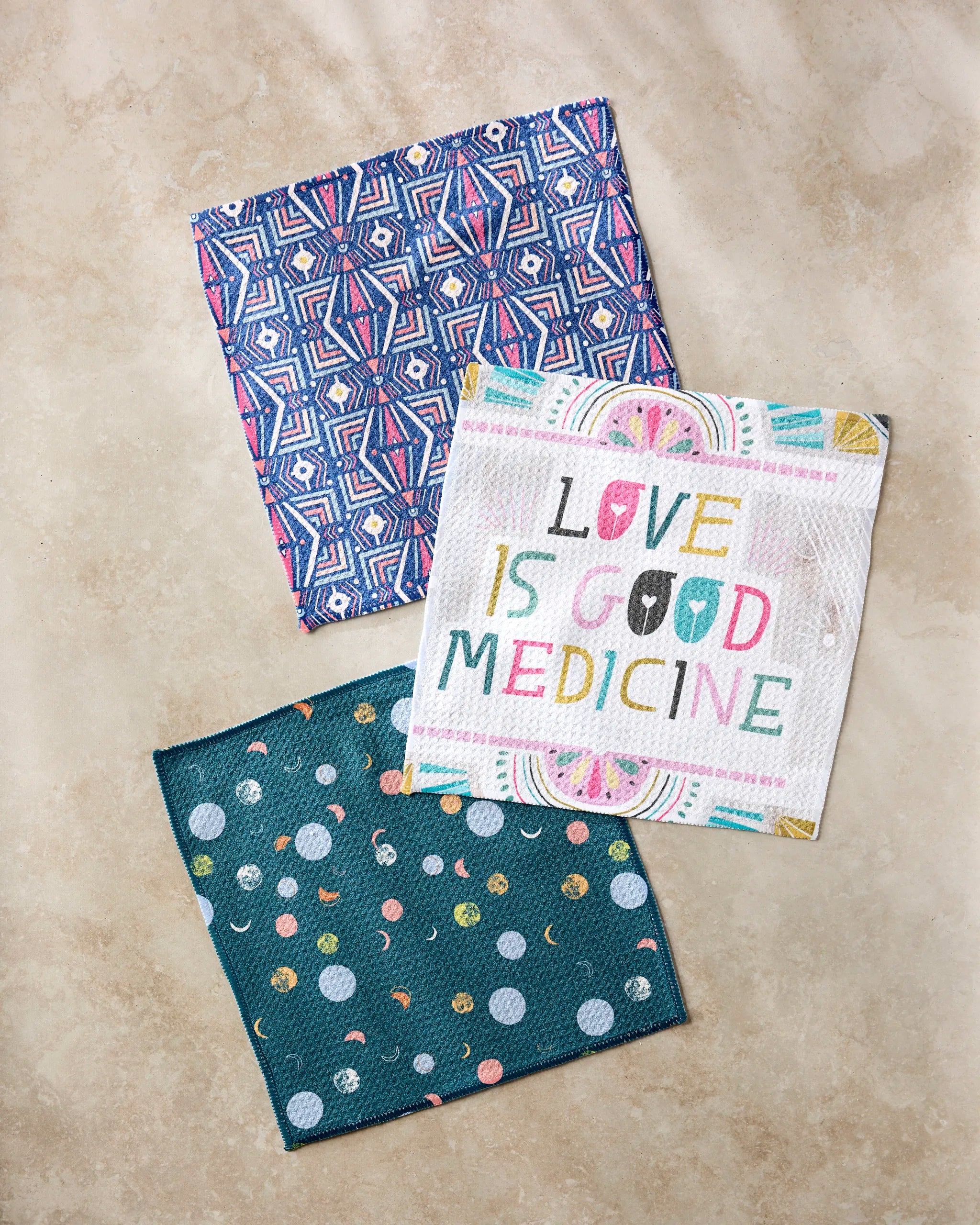 Three patterned cloths from Jessica Swift's "Love is Good Medicine" set sit on a beige surface—featuring a blue geometric towel, a teal one with colorful dots and moons, and a white towel with multicolored text. Made from recycled materials.