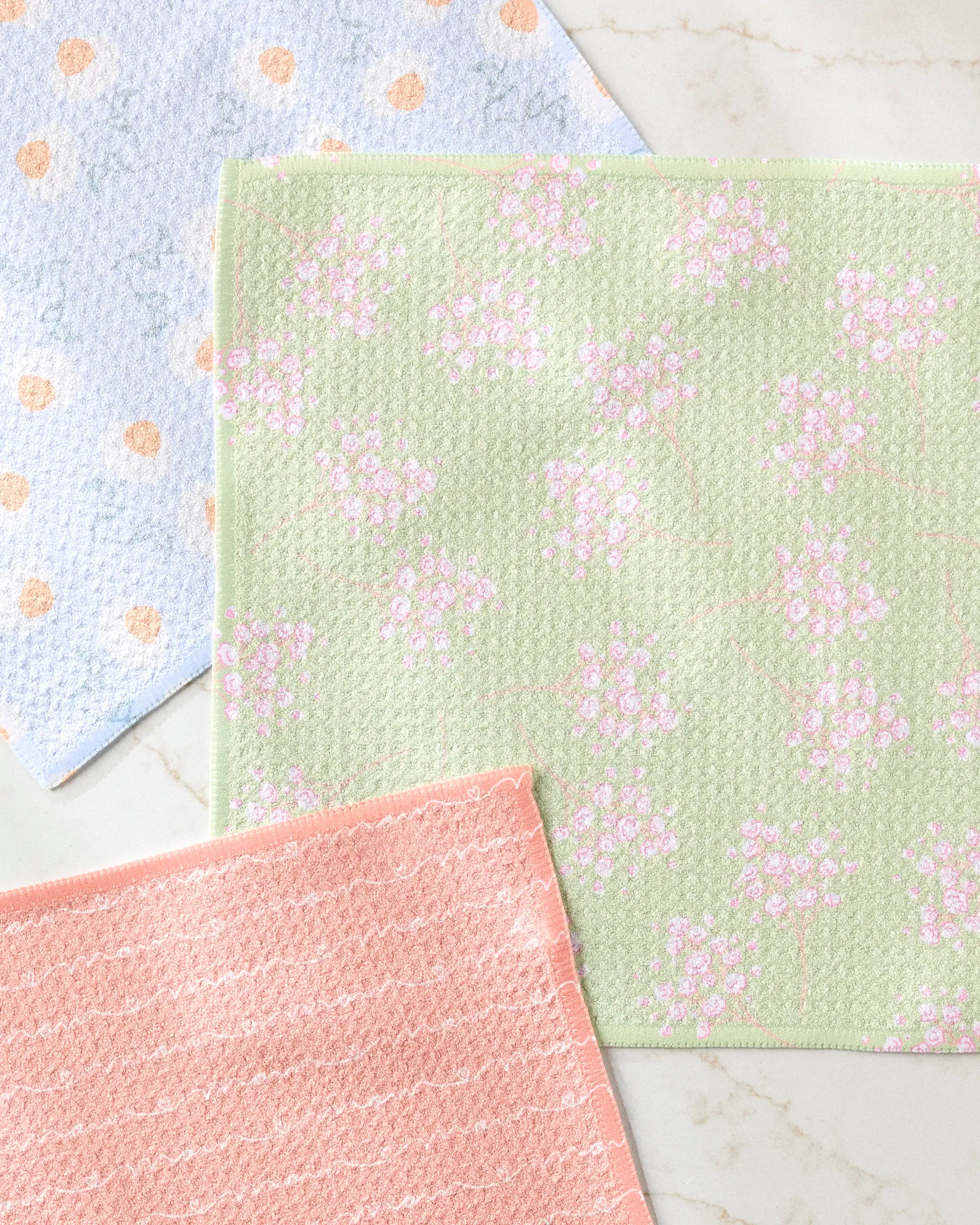 The Love of Mom dishcloth set by Sarah Reed features three absorbent cloths: green with pink florals, peach with wavy lines, and white with blue and orange dots, adding style and color to any kitchen.
