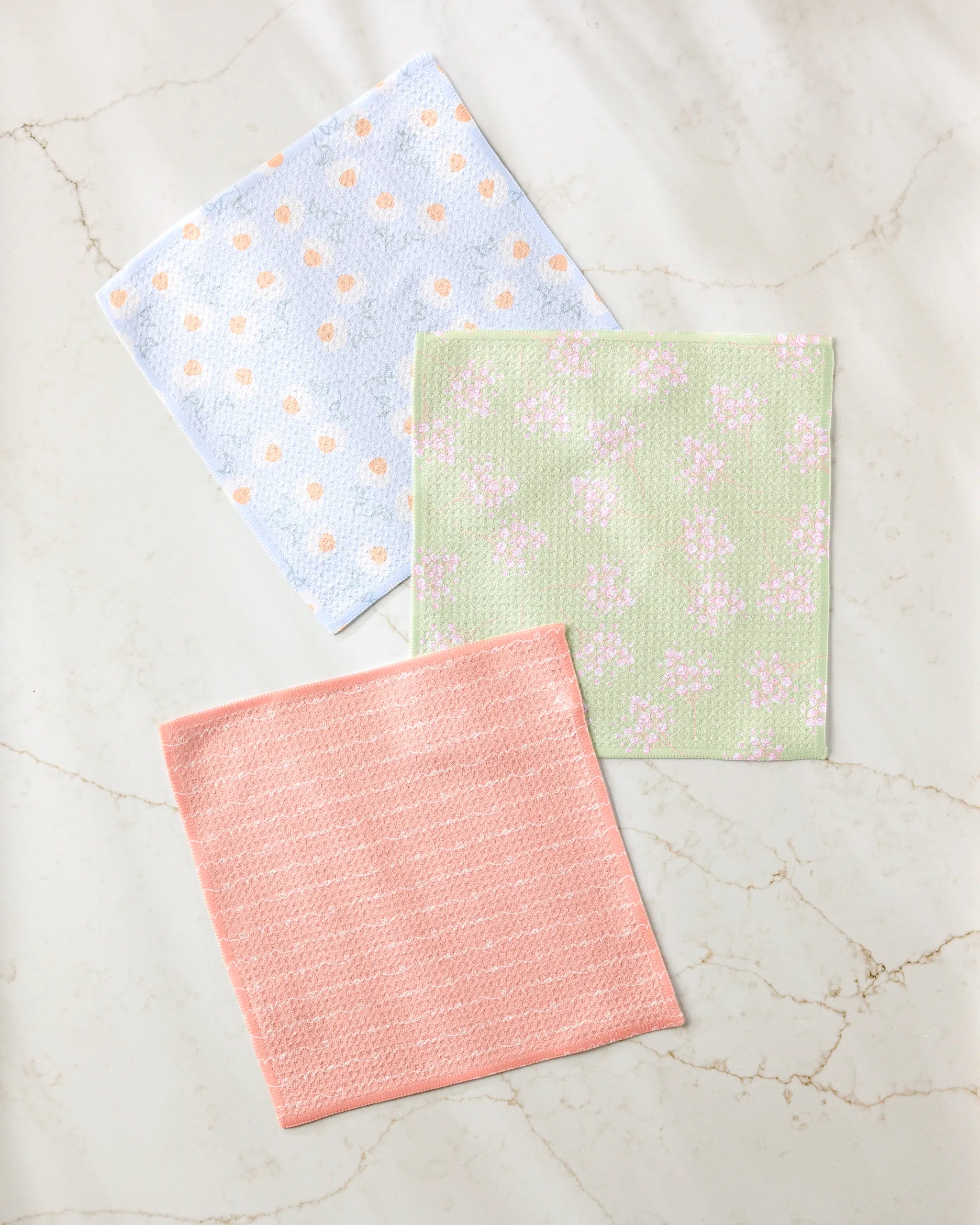 Three square Love of Mom dishcloths by Sarah Reed rest on a marble surface: one blue with orange dots, one green with a white floral design, and one solid pink with a subtle textured pattern.