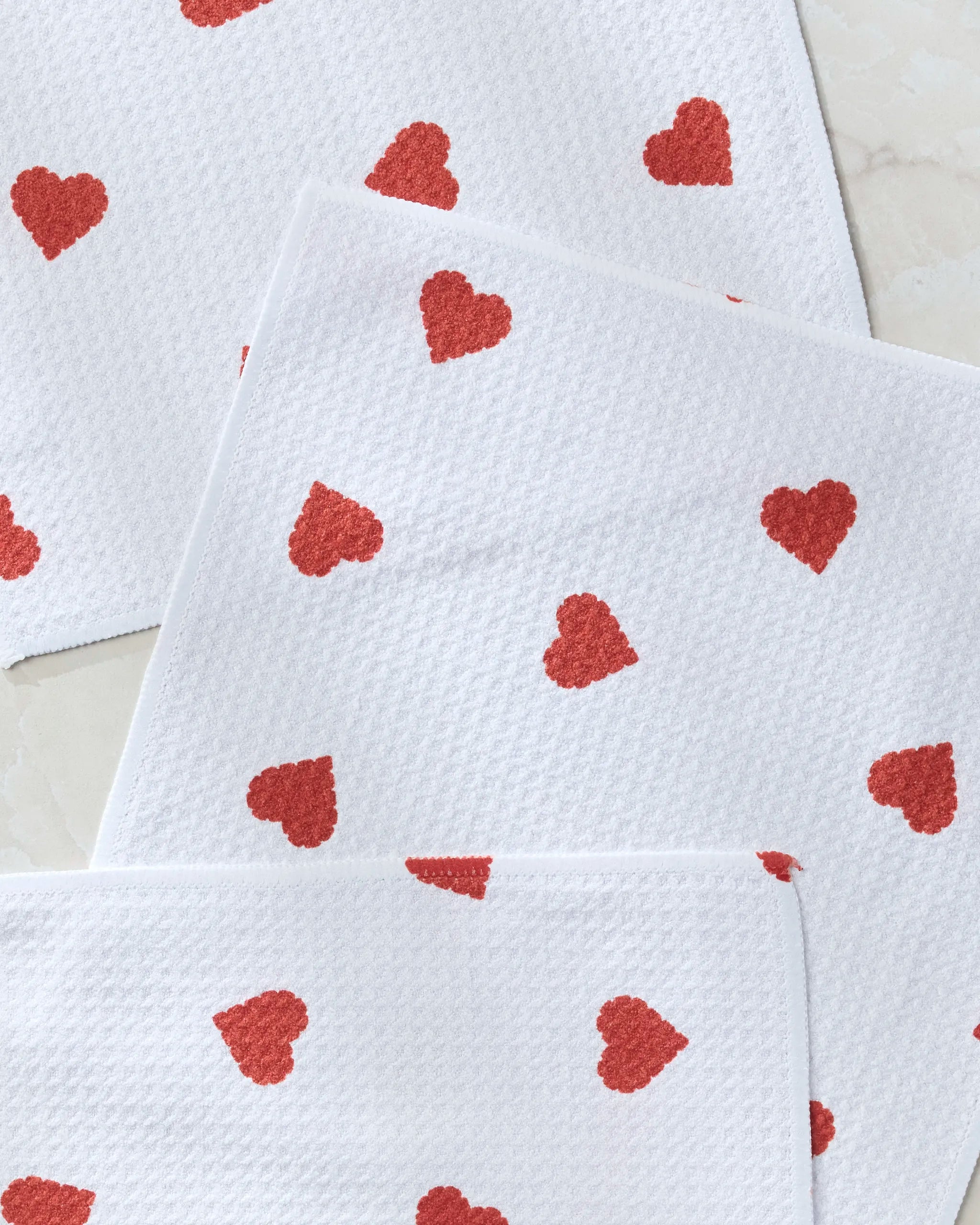 Three “Love Toss” dishtowels by Geometry, featuring white textured fabric with red heart designs, are layered on a light surface.