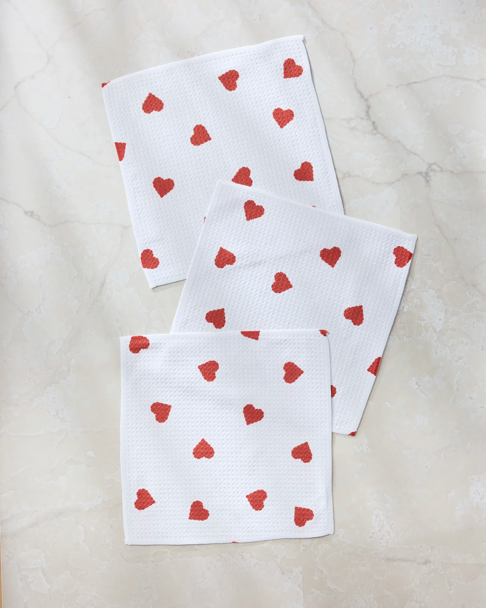 Geometry’s Love Toss dishcloth set features three white napkins adorned with small red hearts, arranged on a marble-like surface—an eco-friendly choice made from recycled materials.