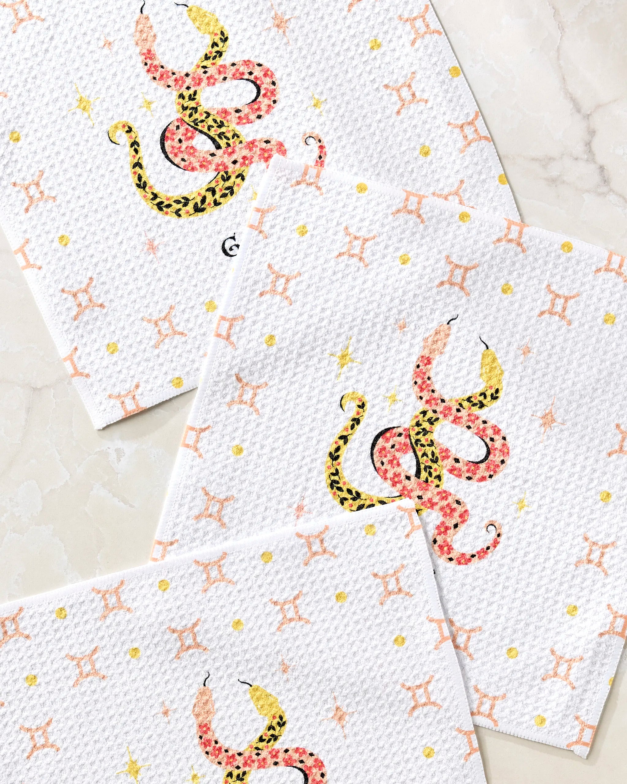 The Lucky Gemini dishcloth set by Heather Zhou features three white recycled cloths with orange stars and a bold pink, yellow, and black floral snake design, beautifully displayed on a light marble surface.