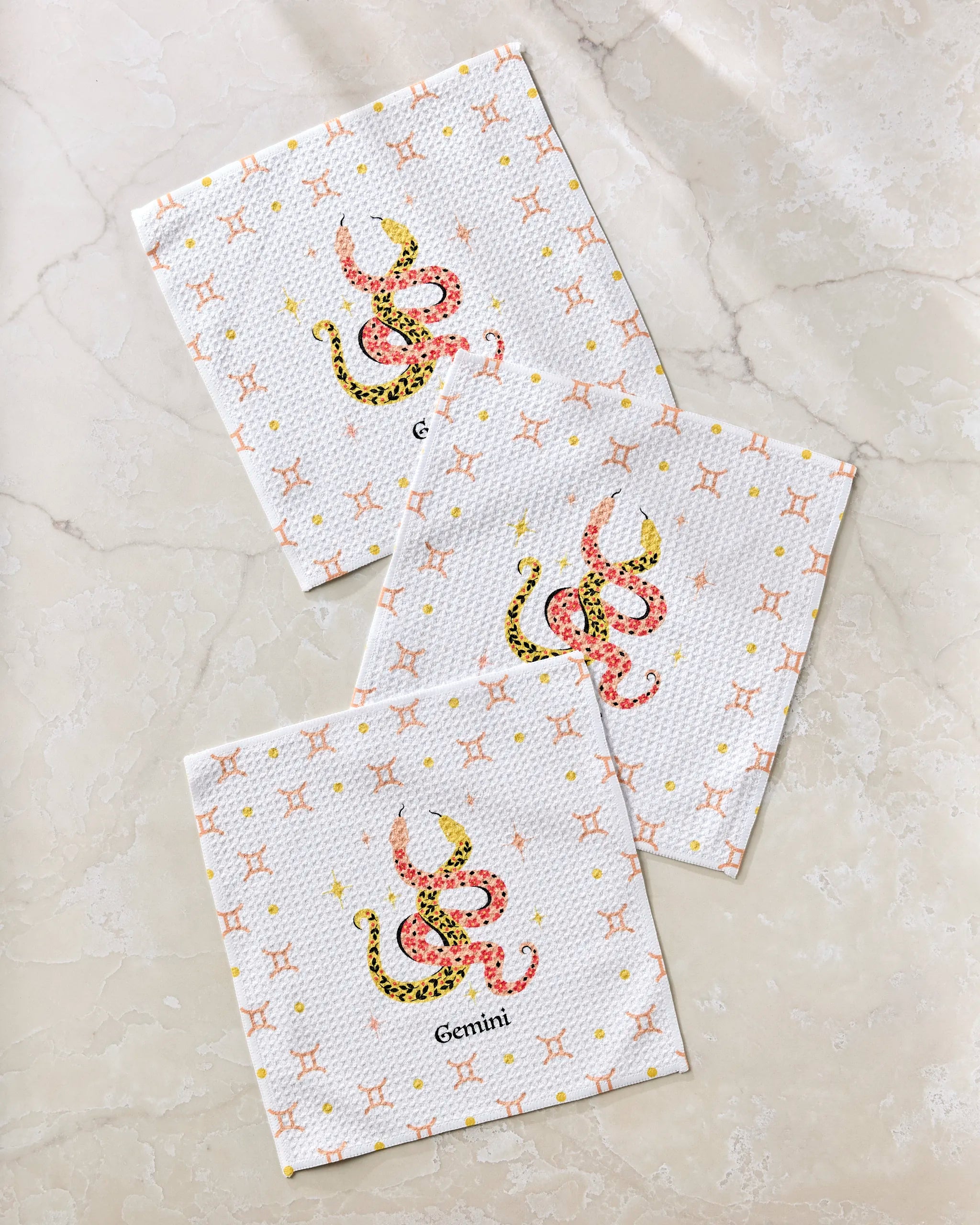 Heather Zhou's Lucky Gemini set includes three white recycled dishcloths featuring a pink and yellow Gemini zodiac symbol, “Gemini” text, and a subtle geometric pattern, all arranged on a light marble surface.