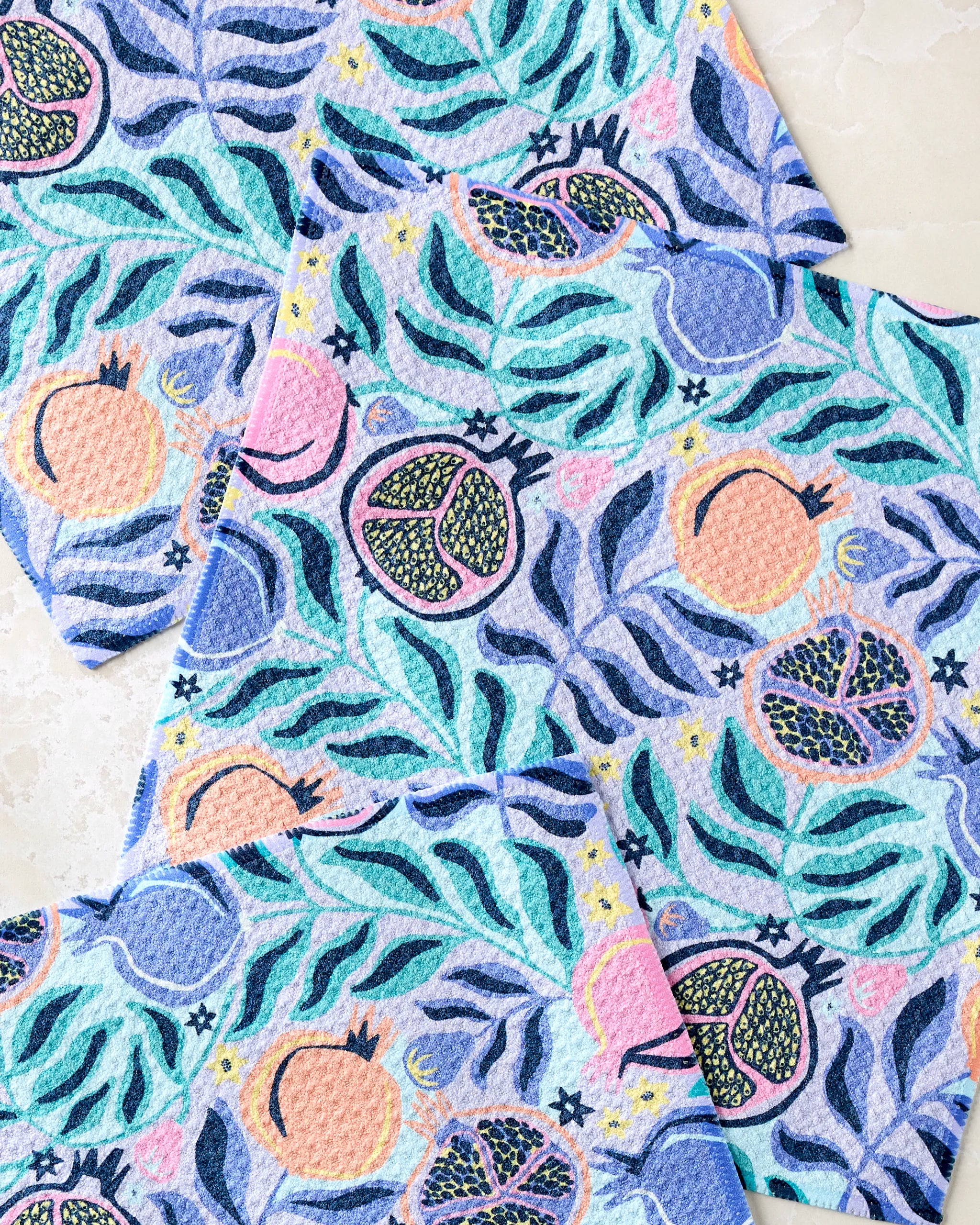 The Lucky Pomegranate dishcloth set by Masha Volnova features a vibrant blue, green, pink, and orange pomegranate and leaf pattern on super-absorbent recycled material dishtowels.