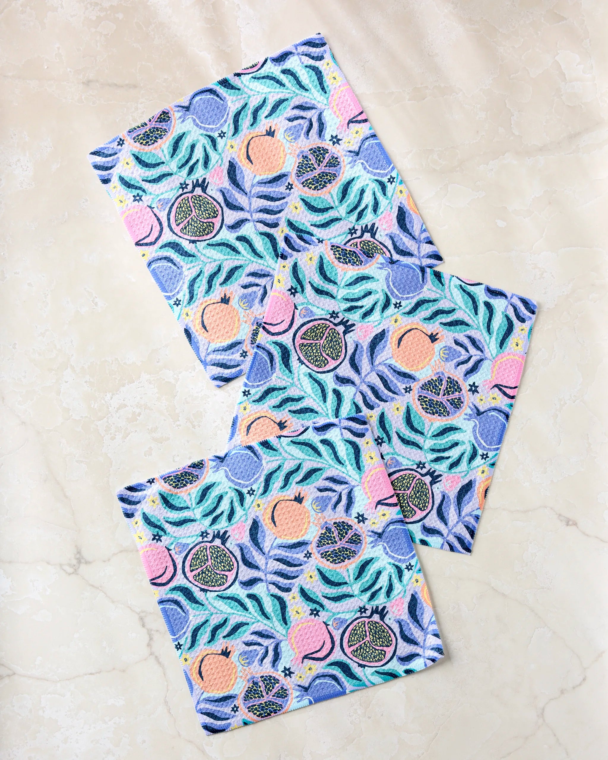 Three colorful, super-absorbent Lucky Pomegranate dishtowels by Masha Volnova, featuring vibrant fig, peach, and leaf patterns in blue, green, pink, and purple, are arranged on a light marble surface.