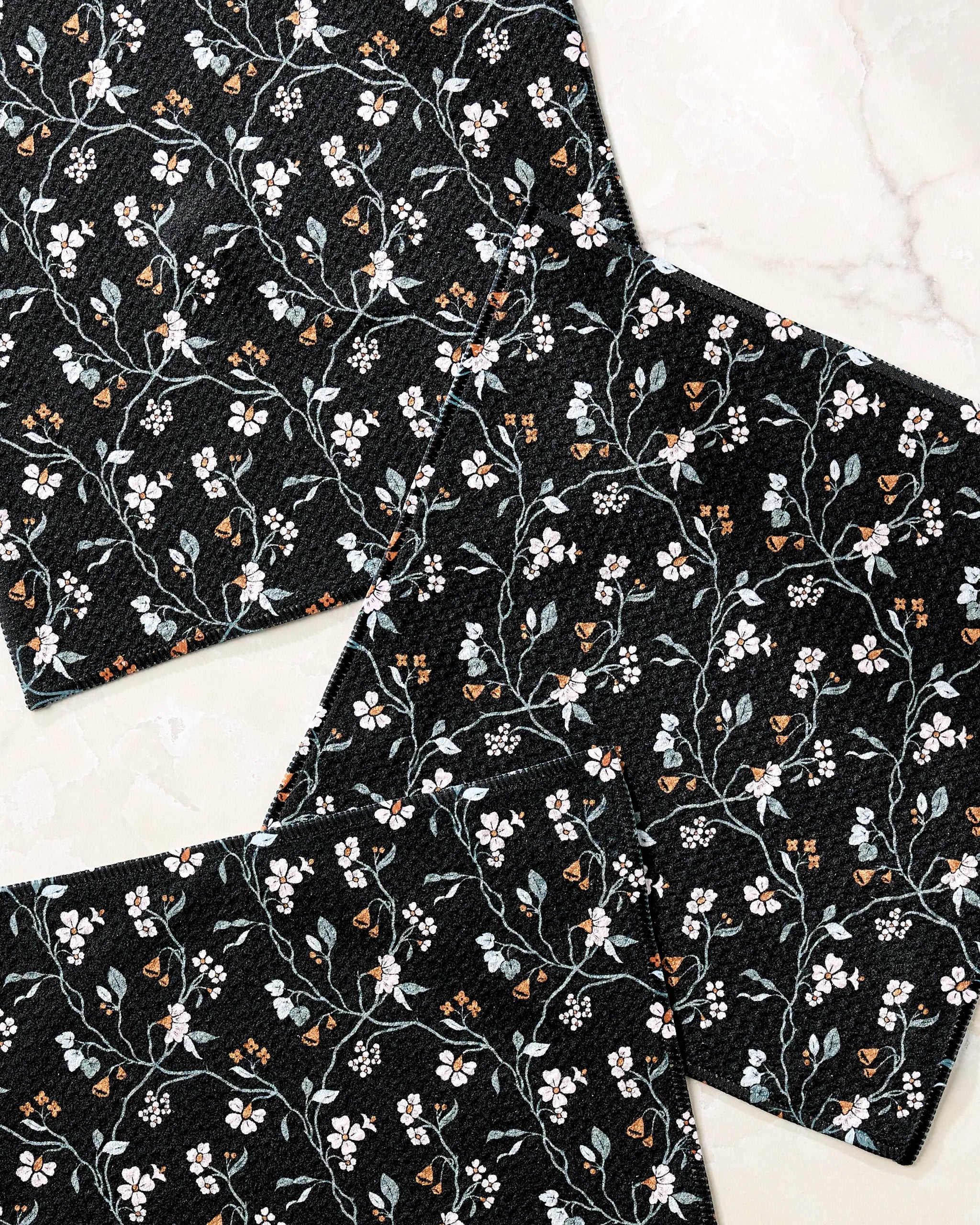 Four Lunaria in Charcoal placemats by Danelys Sidron, featuring a white and light blue floral pattern, arranged on a marble surface, pair beautifully with recycled dishcloths for an eco-friendly dining space.