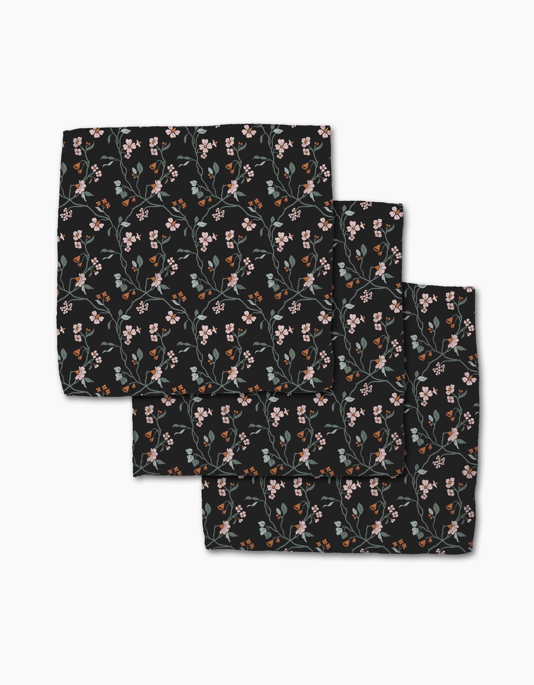 The Lunaria in Charcoal napkins by Danelys Sidron are three neatly stacked black cloth napkins, made from recycled materials, featuring a pattern of small pink, white, and orange flowers with green stems and leaves.