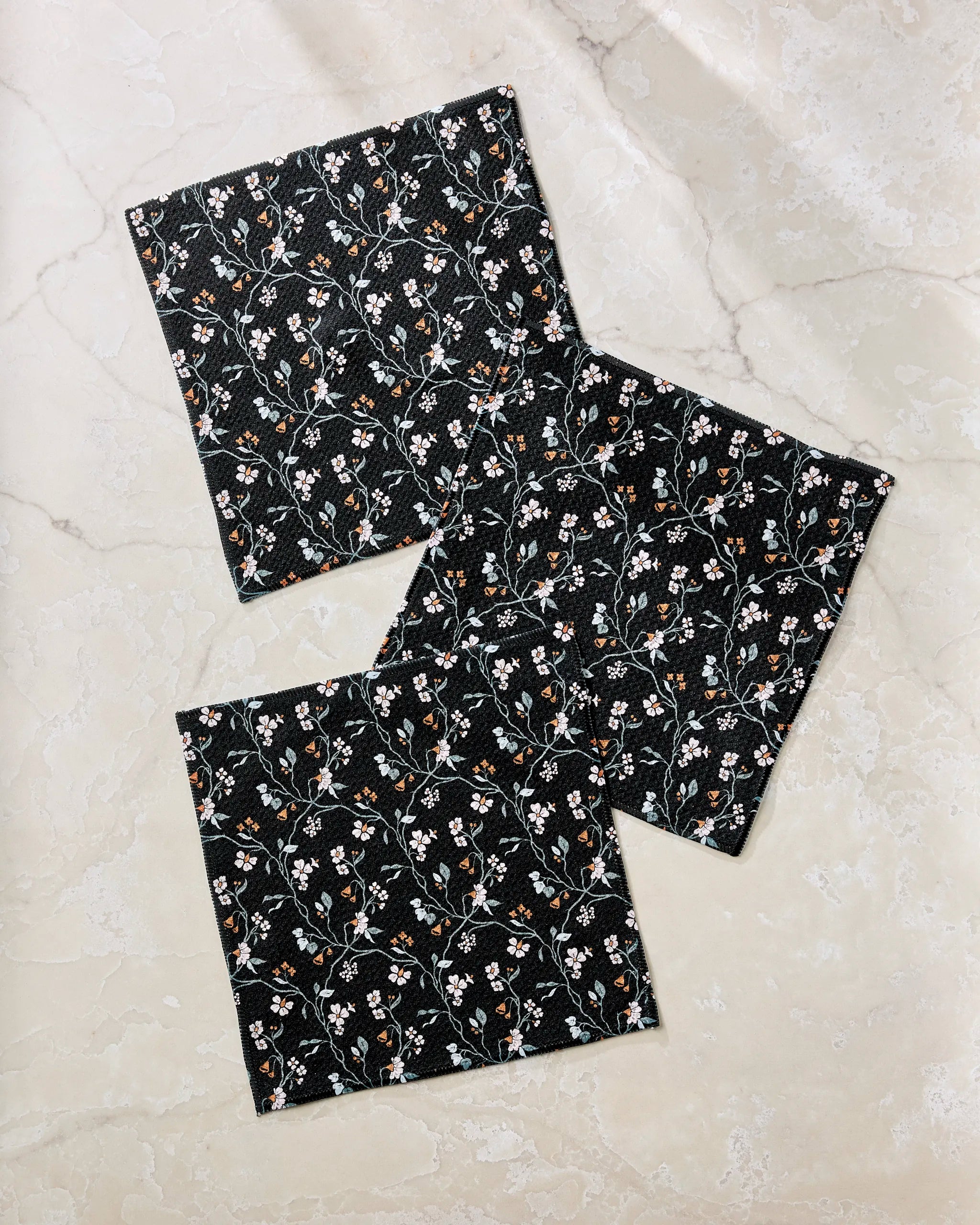 Three square Lunaria in Charcoal dishcloths by Danelys Sidron, featuring a white and orange floral pattern on black fabric, are arranged diagonally on a light marble surface. Crafted from recycled materials.