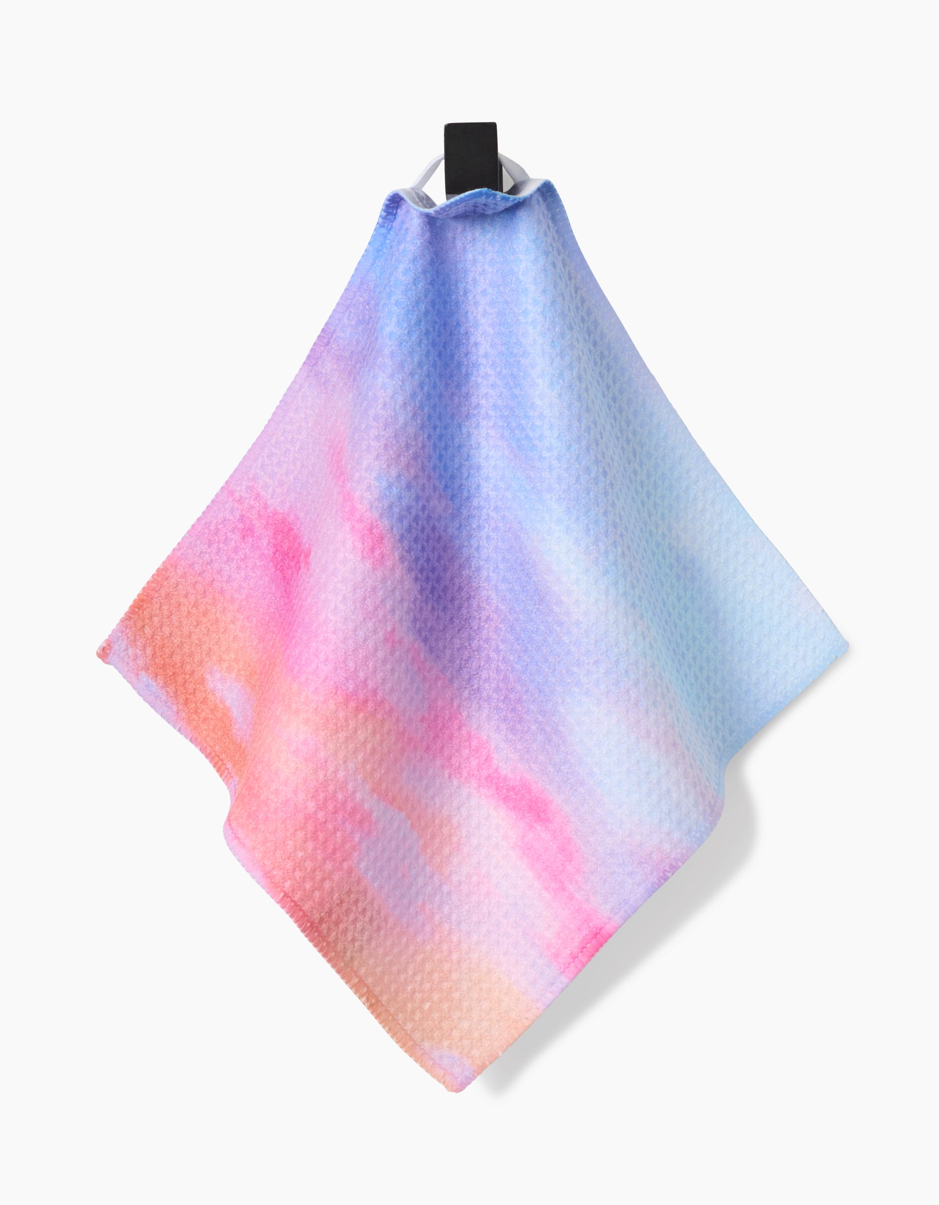 The Magic In The Sky kitchen towel by Stefanie Bales, crafted from recycled materials with a waffle texture in pastel pink, blue, and orange tones, hangs from a black hook against a plain white background.