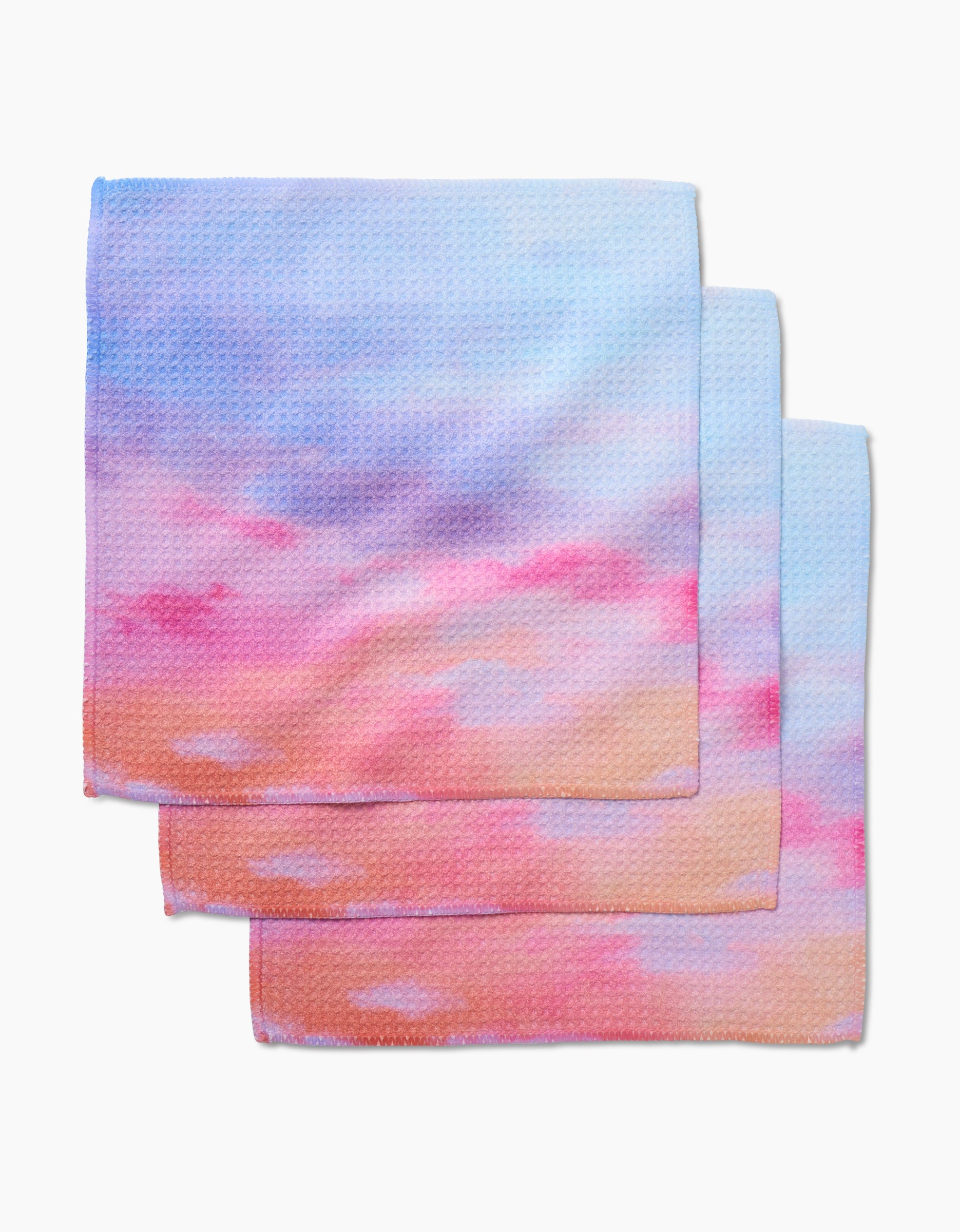 Three Magic In The Sky dishcloths by Stefanie Bales, made from Geoweave fabric and featuring a soft blue, pink, and orange pastel gradient, are layered slightly atop each other on a white background.