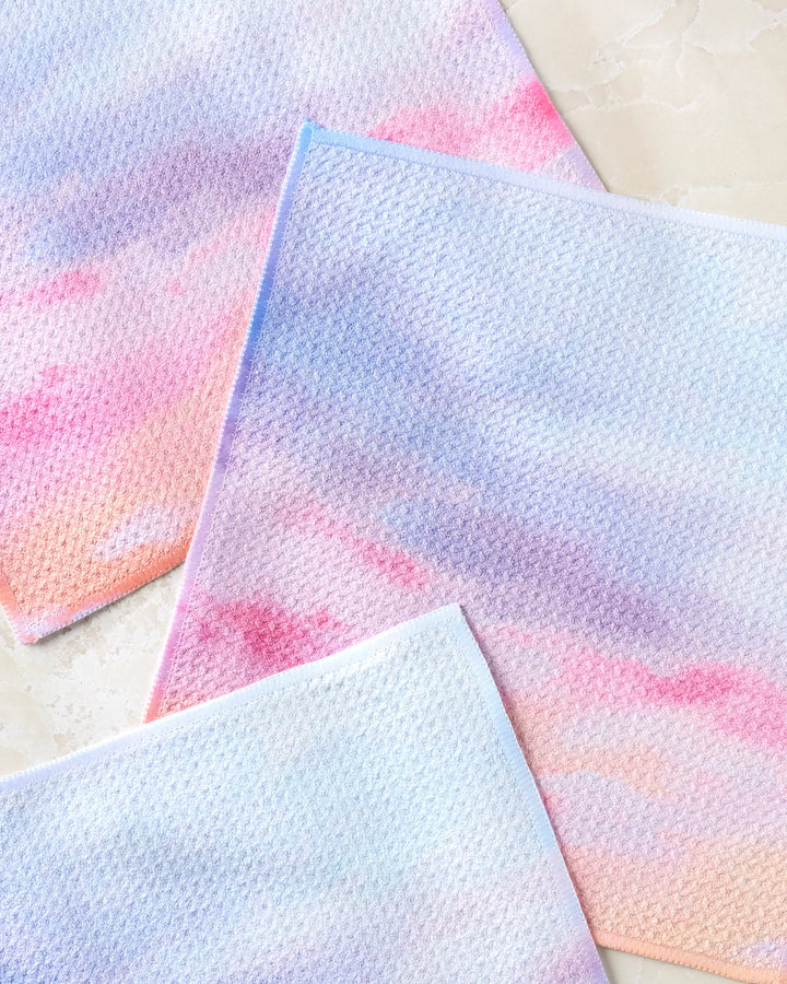 Three super-absorbent "Magic In The Sky" dishtowels by Stefanie Bales, featuring a pastel gradient in pink, purple, and blue, are displayed on a marble-like surface—combining charm and efficiency for your kitchen.