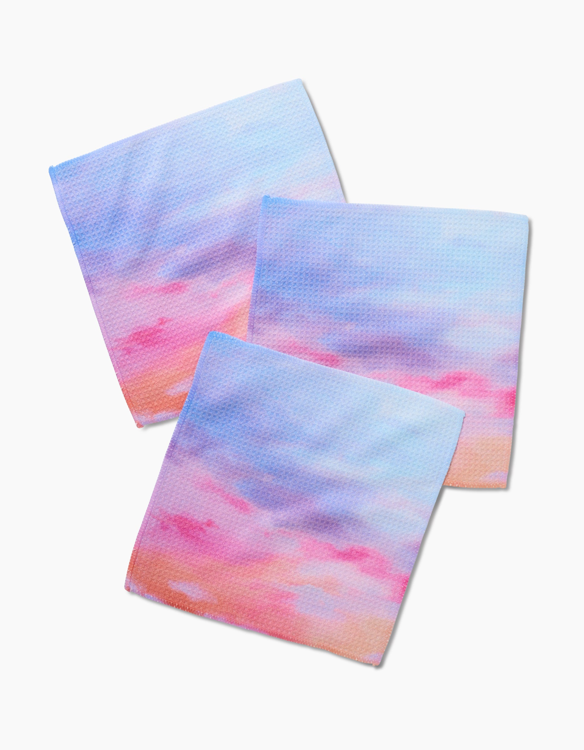 Three Magic In The Sky Geoweave dishcloths by Stefanie Bales, featuring a pastel sunset-inspired gradient in blue, pink, and purple, are arranged overlapping on a plain white background.