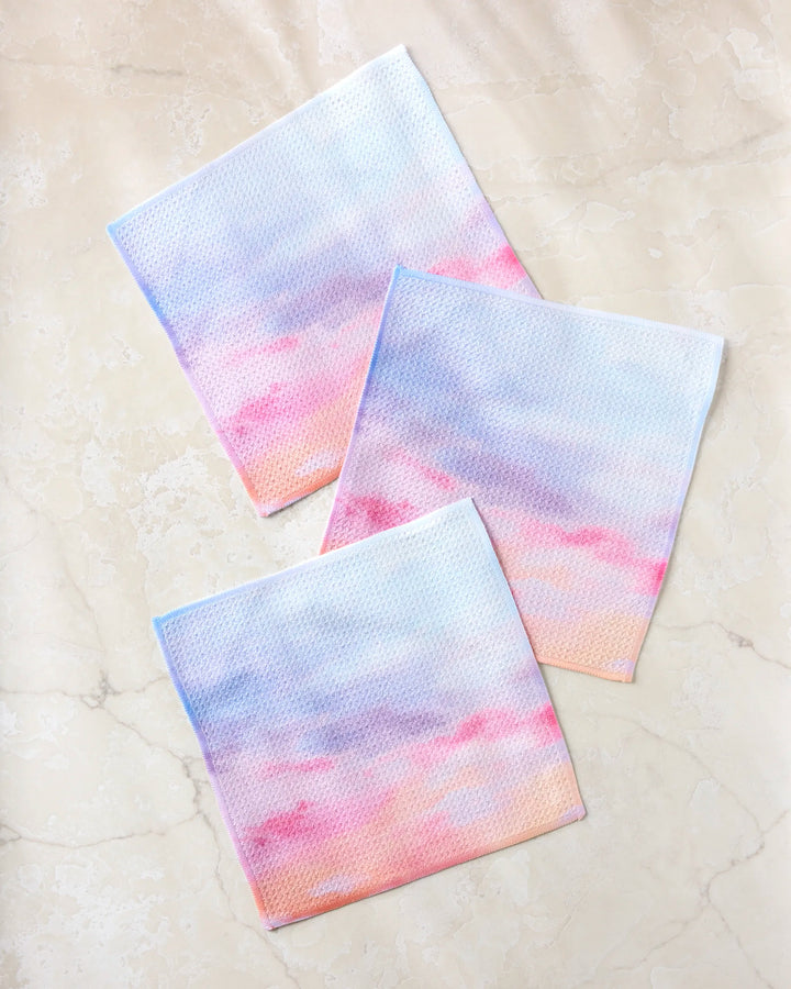 Three "Magic In The Sky" dishtowels by Stefanie Bales, in pastel blue, pink, purple, and peach gradients, are displayed on a light marble surface.