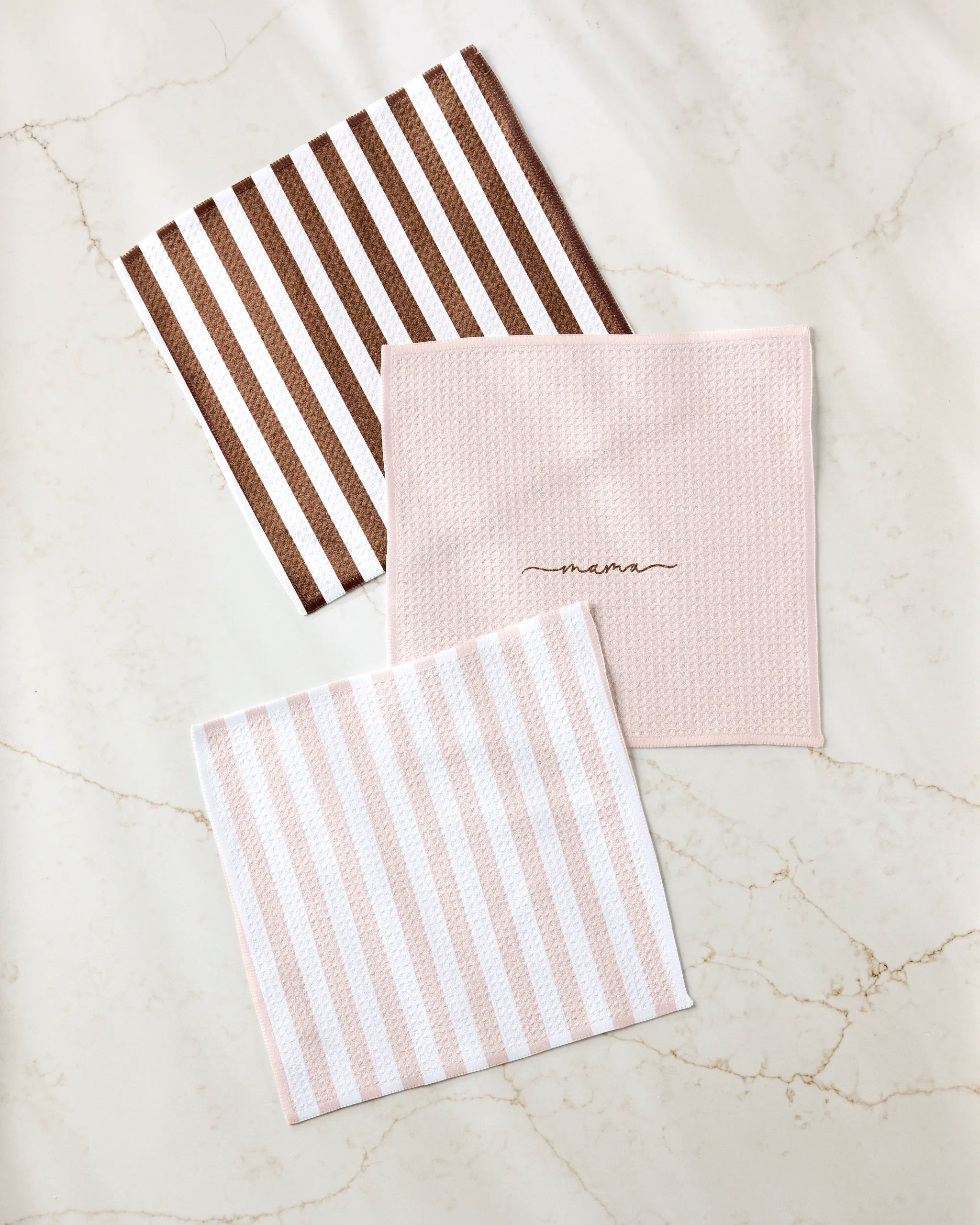 Three folded Kailee Wright "Mama" cloth napkins rest on marble: one with brown and white stripes, one solid light pink with "mama" in cursive, and one with light pink and white stripes, all made from recycled materials.