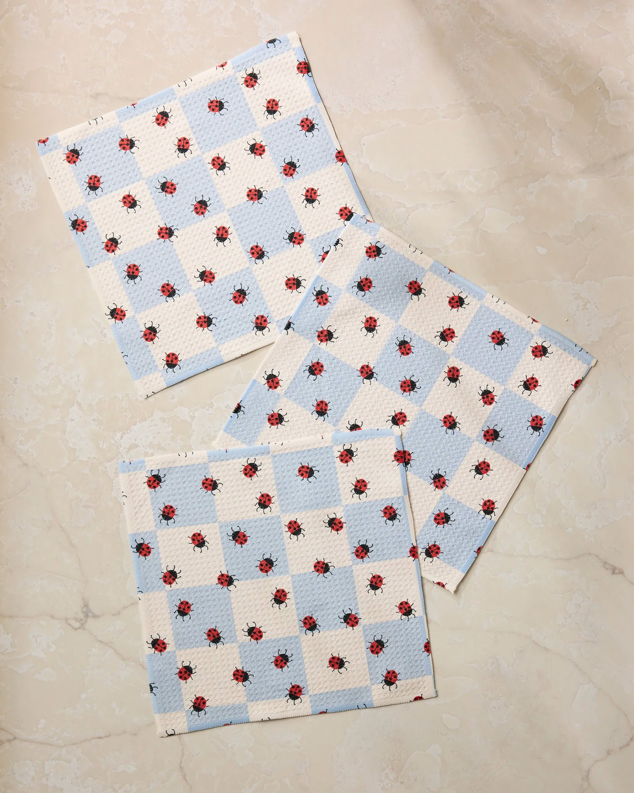 Three square cloths from Geometry's Mariquita Pop set, featuring a blue and white checkered pattern with small red ladybugs, overlap on a marble surface—a charming dishcloth set made from recycled materials.