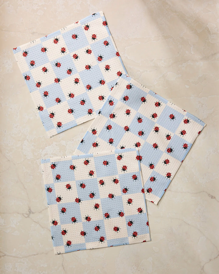 Three square cloths from Geometry's Mariquita Pop set, featuring a blue and white checkered pattern with small red ladybugs, overlap on a marble surface—a charming dishcloth set made from recycled materials.