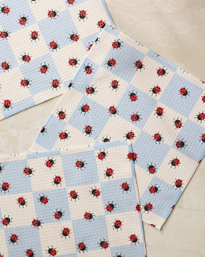 Four Geometry Mariquita Pop placemats, featuring a blue and white checkered pattern with red ladybugs, are arranged on a beige marbled surface.