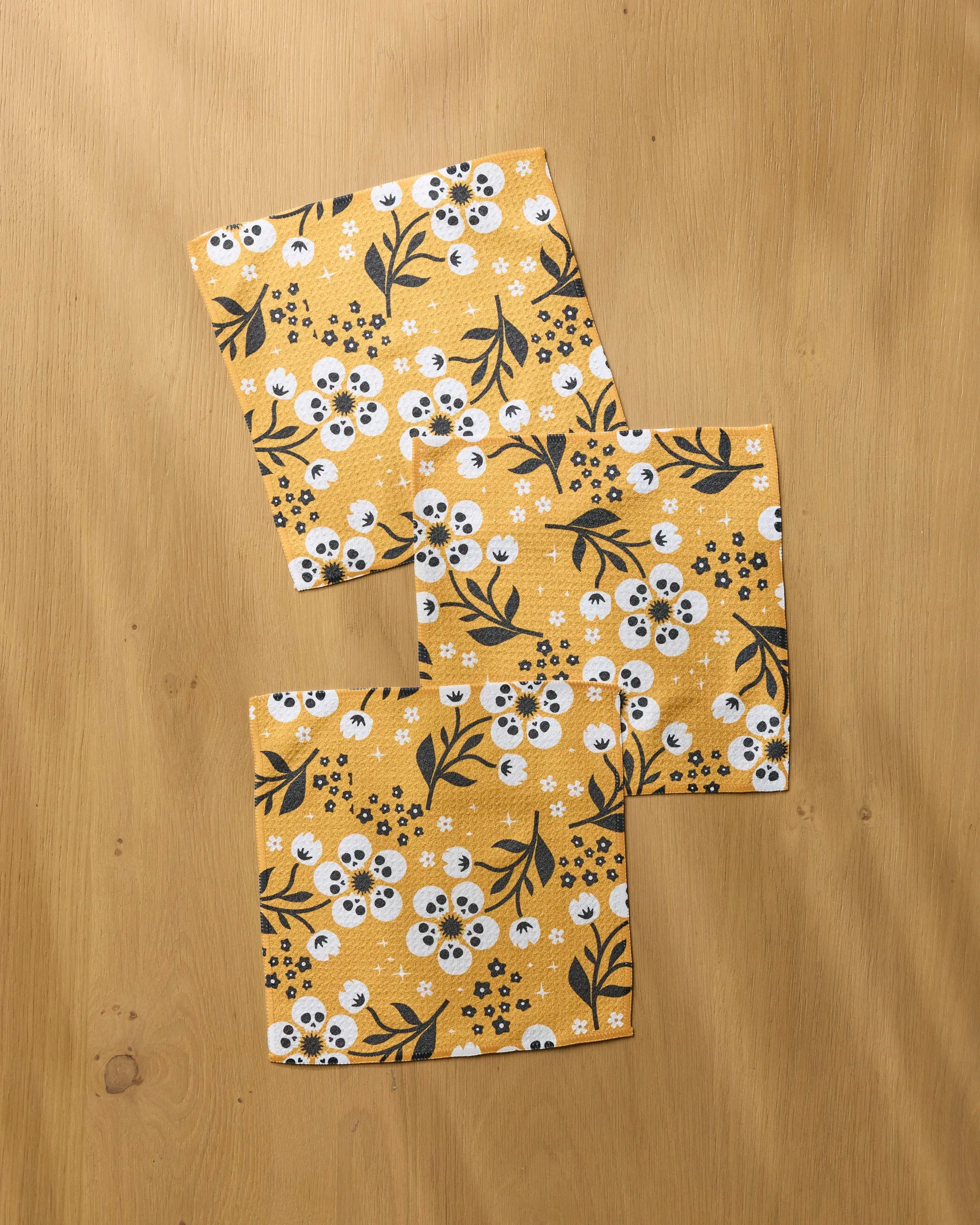 Three "Marrow Bloom" cloth napkins by Geometry, featuring a yellow base with a white and black floral design, are stylishly arranged on a light wooden surface—ideal for use as a reusable dishcloth set.