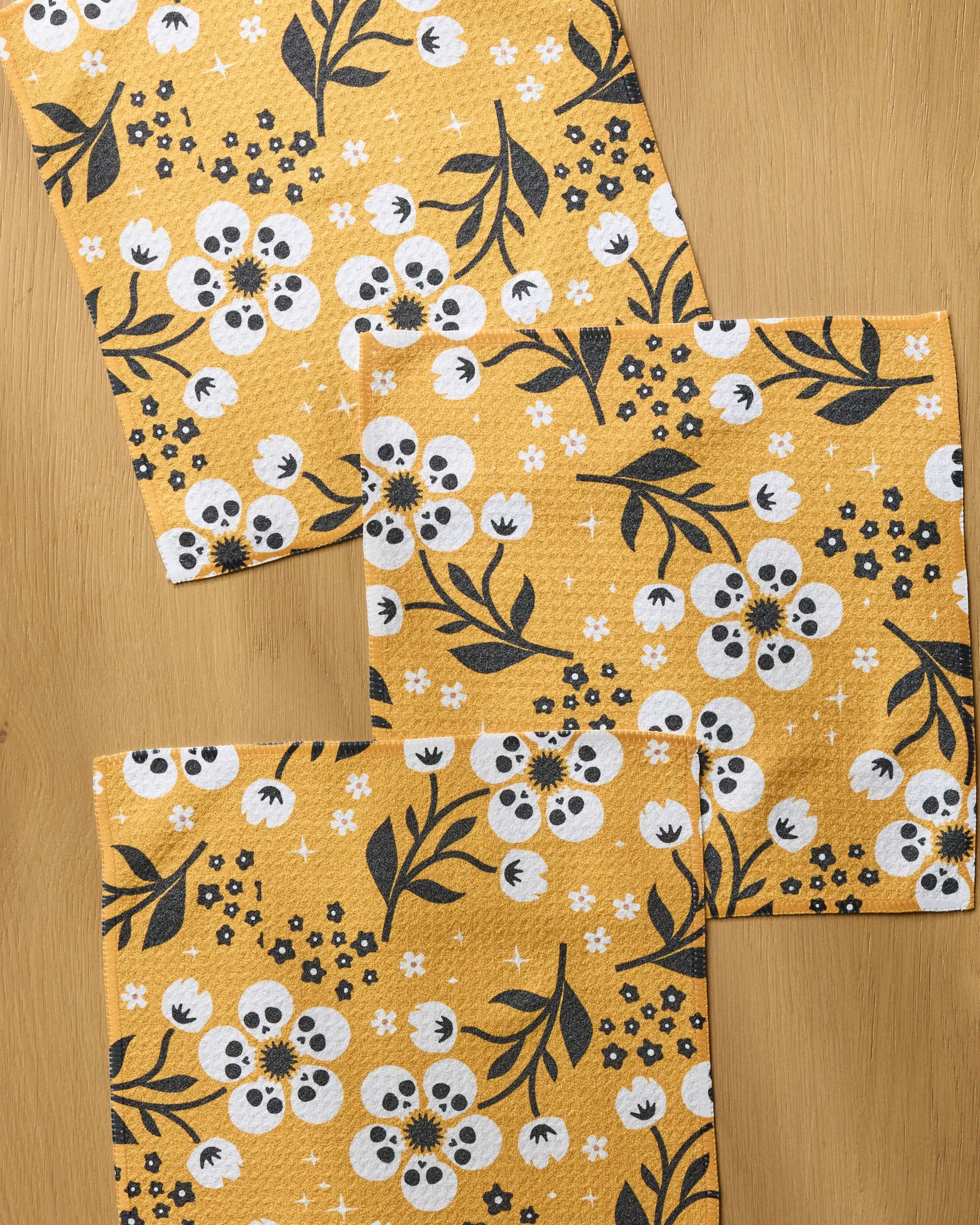 Three Marrow Bloom reusable dishcloths by Geometry, featuring a white and black floral pattern with flowers, leaves, and star shapes on a yellow background, are partly overlapping on a light wooden surface.