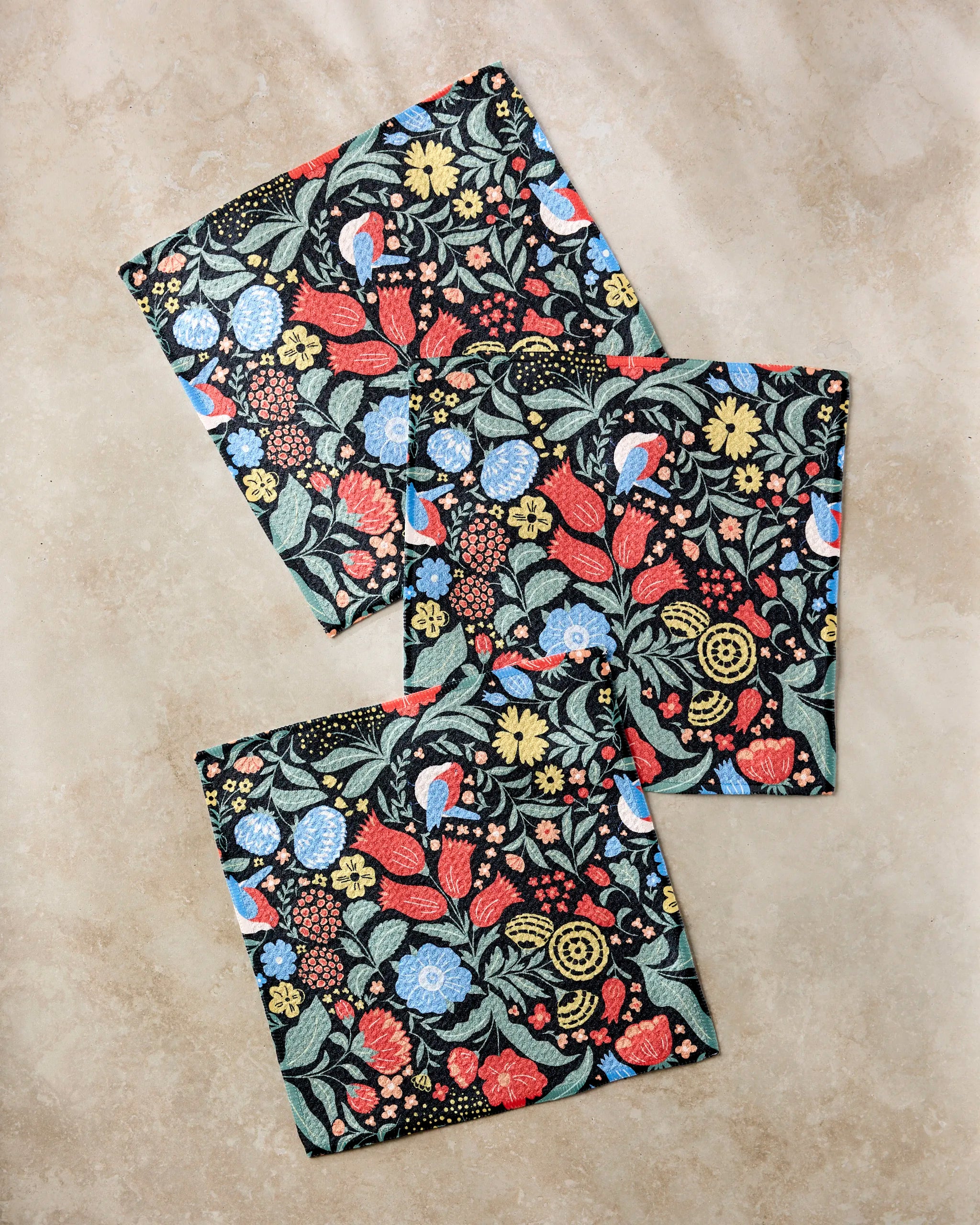 The Marsh Wren napkins by Danelys Sidron are square and made from recycled materials, featuring a colorful floral and bird pattern with red, blue, and yellow flowers, green leaves, and blue birds.