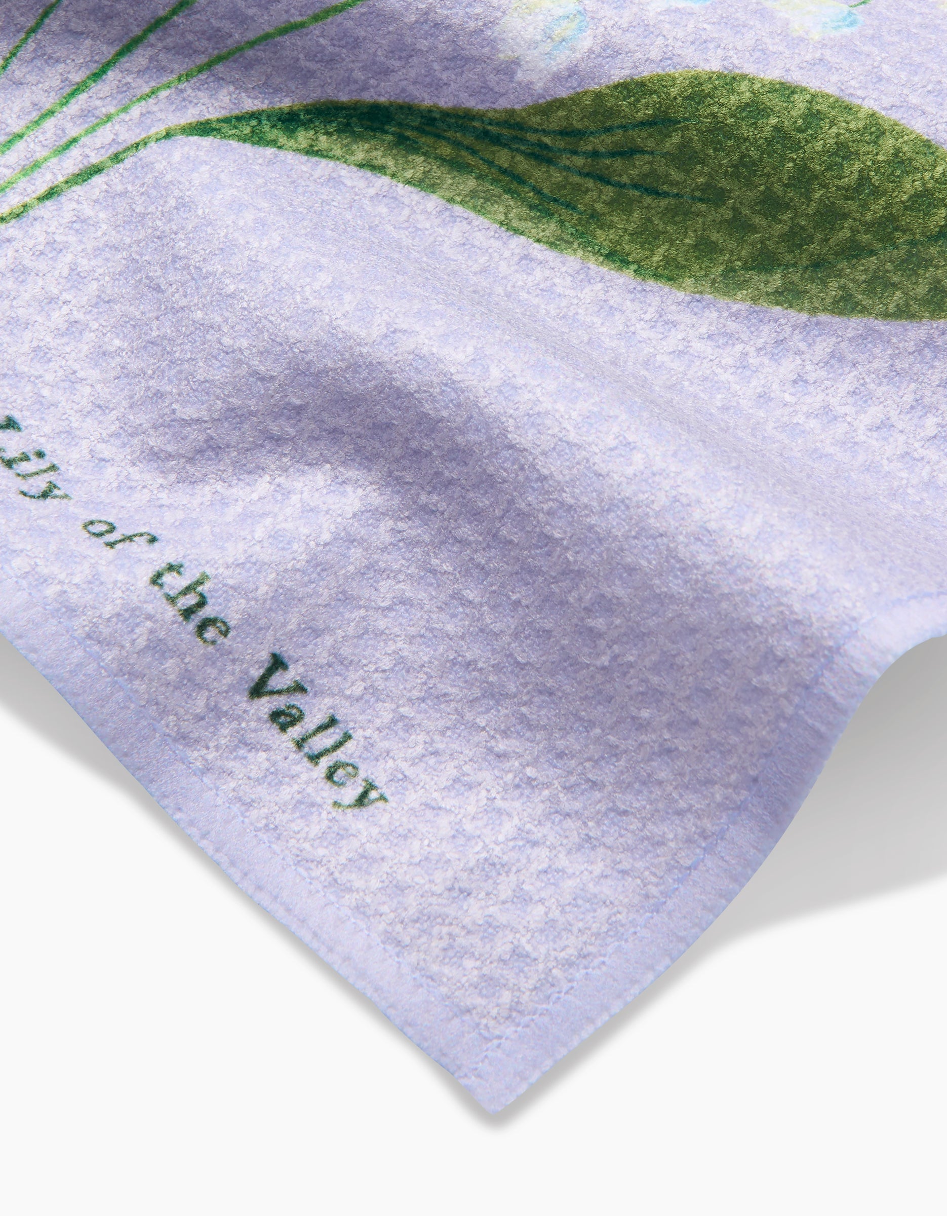 A close-up of the textured lavender May Lilly Of The Valley dishcloth by Linda Fox, featuring a partial green leaf design and Lily of the Valley text along the edge.