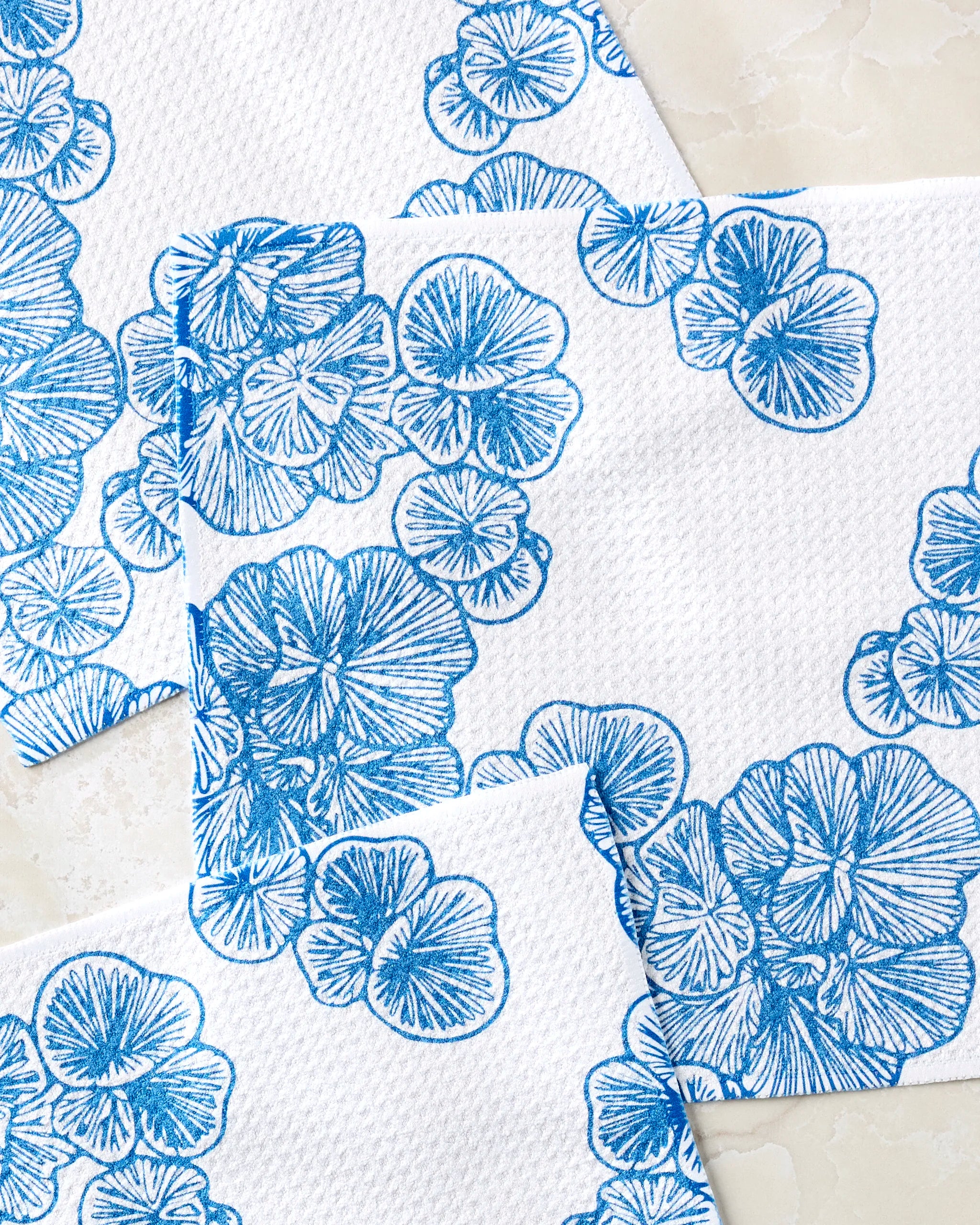 Meadowline napkins by Second Nature Studio, made from recycled materials and featuring blue floral patterns, are stylishly overlapped on a light marble surface.