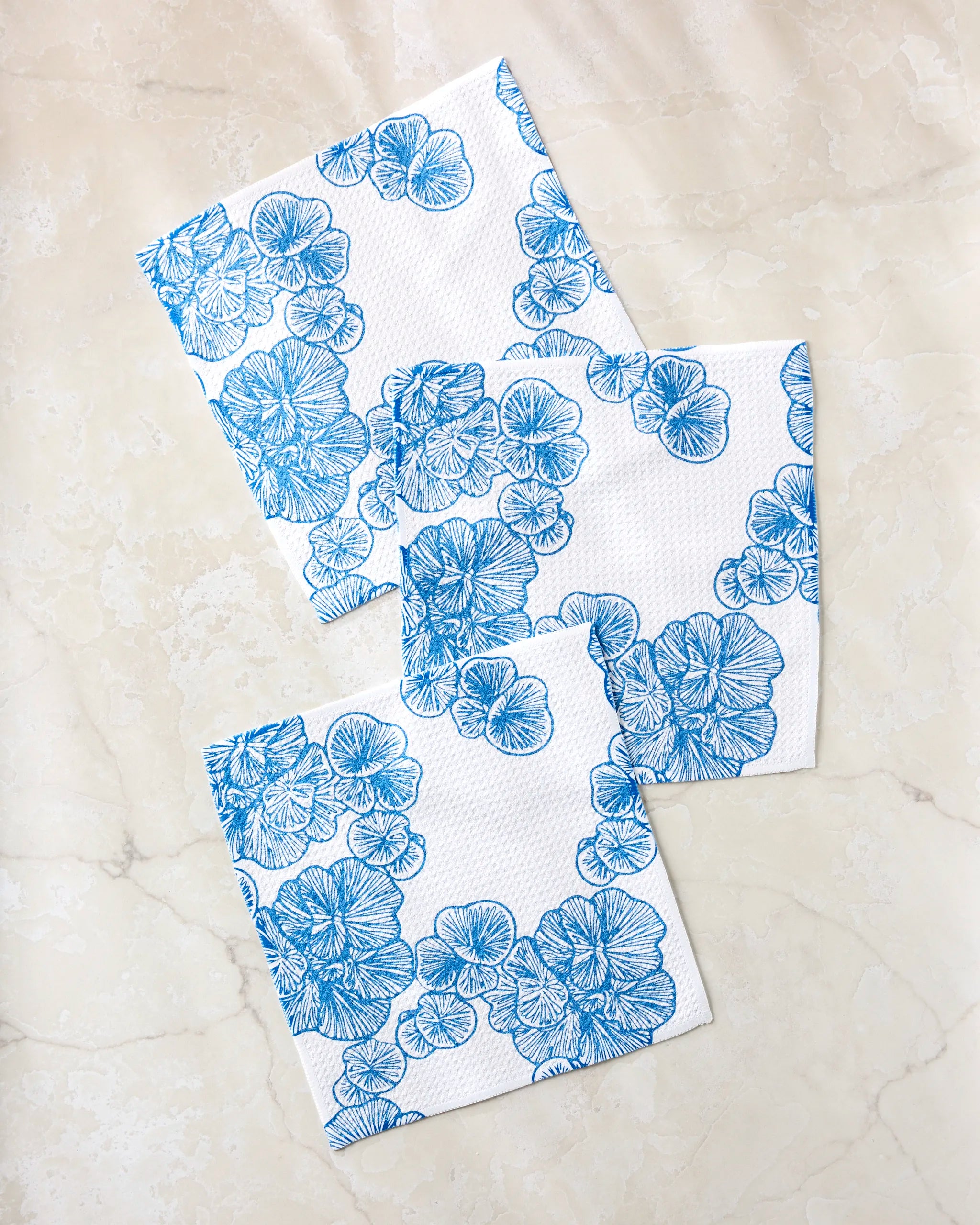 Three Meadowline napkins by Second Nature Studio, featuring blue floral patterns on white and made from recycled materials, are arranged with overlapping corners on a light marble surface.