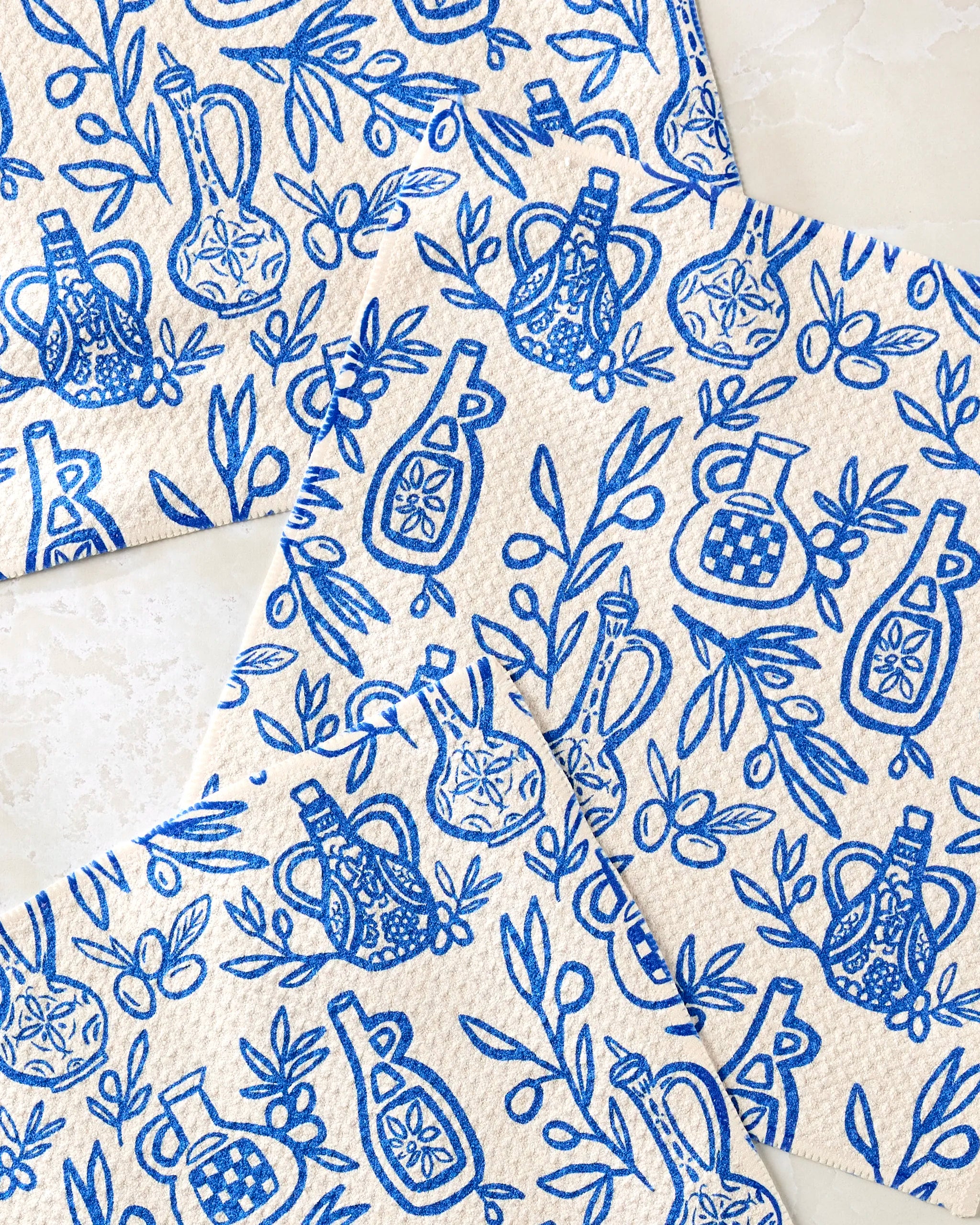 The "Mediterranean Pour" dishcloth set by Lisa Nelson features blue and white patterns of olive oil bottles, branches, and olives on a marble background. Made from recycled kitchen cloths for style and sustainability.
