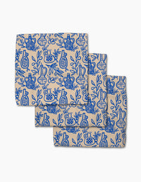 Three Mediterranean Pour beige cloth napkins by Lisa Nelson, crafted from recycled materials and featuring a blue line art pattern of vases, pomegranates, and leafy branches, shown overlapping on a white background.