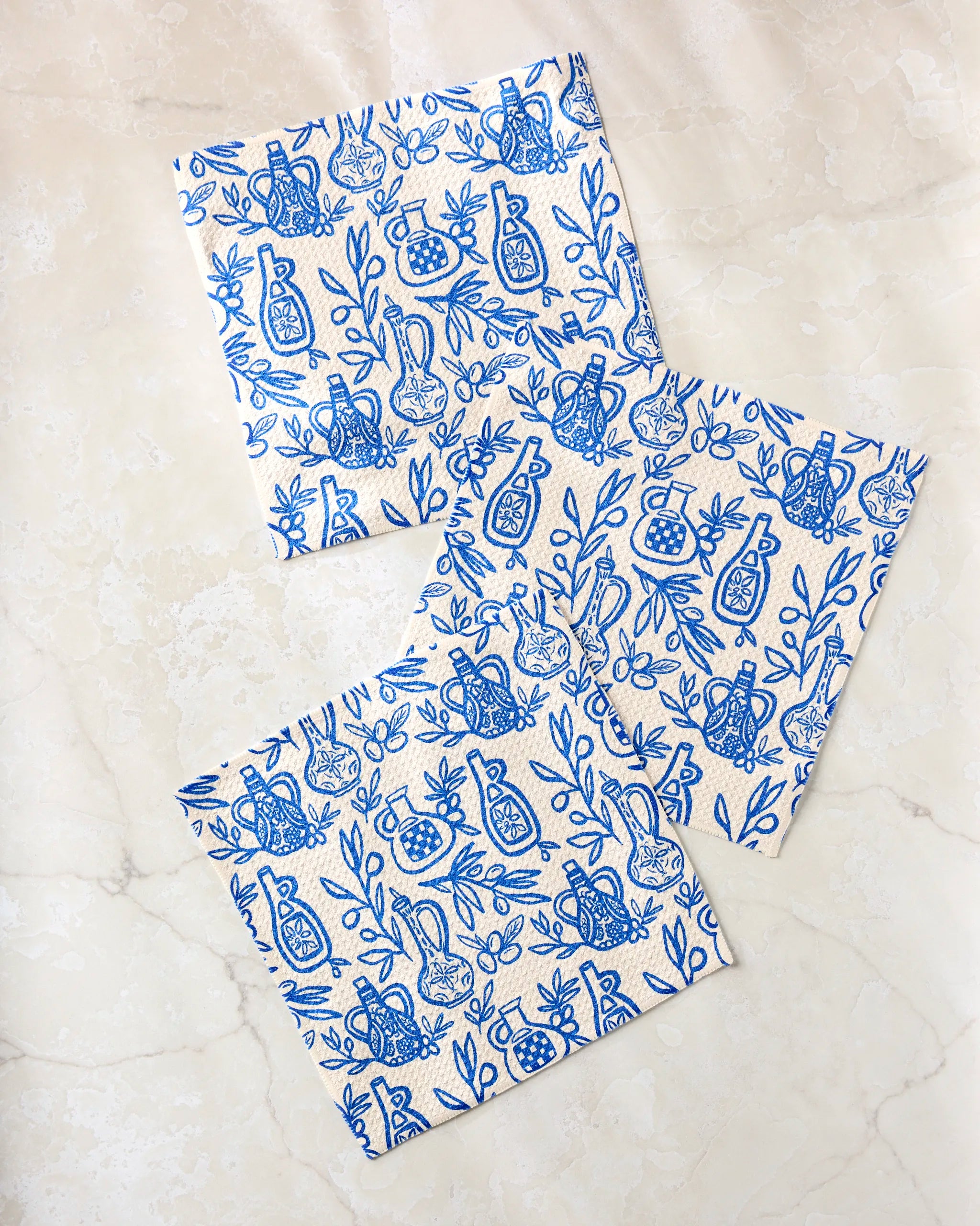 Three "Mediterranean Pour" square napkins by Lisa Nelson, made from recycled kitchen cloths, display a blue and white pattern of vases, pomegranates, and leaves. The napkins are scattered and overlapping on a light marble surface.