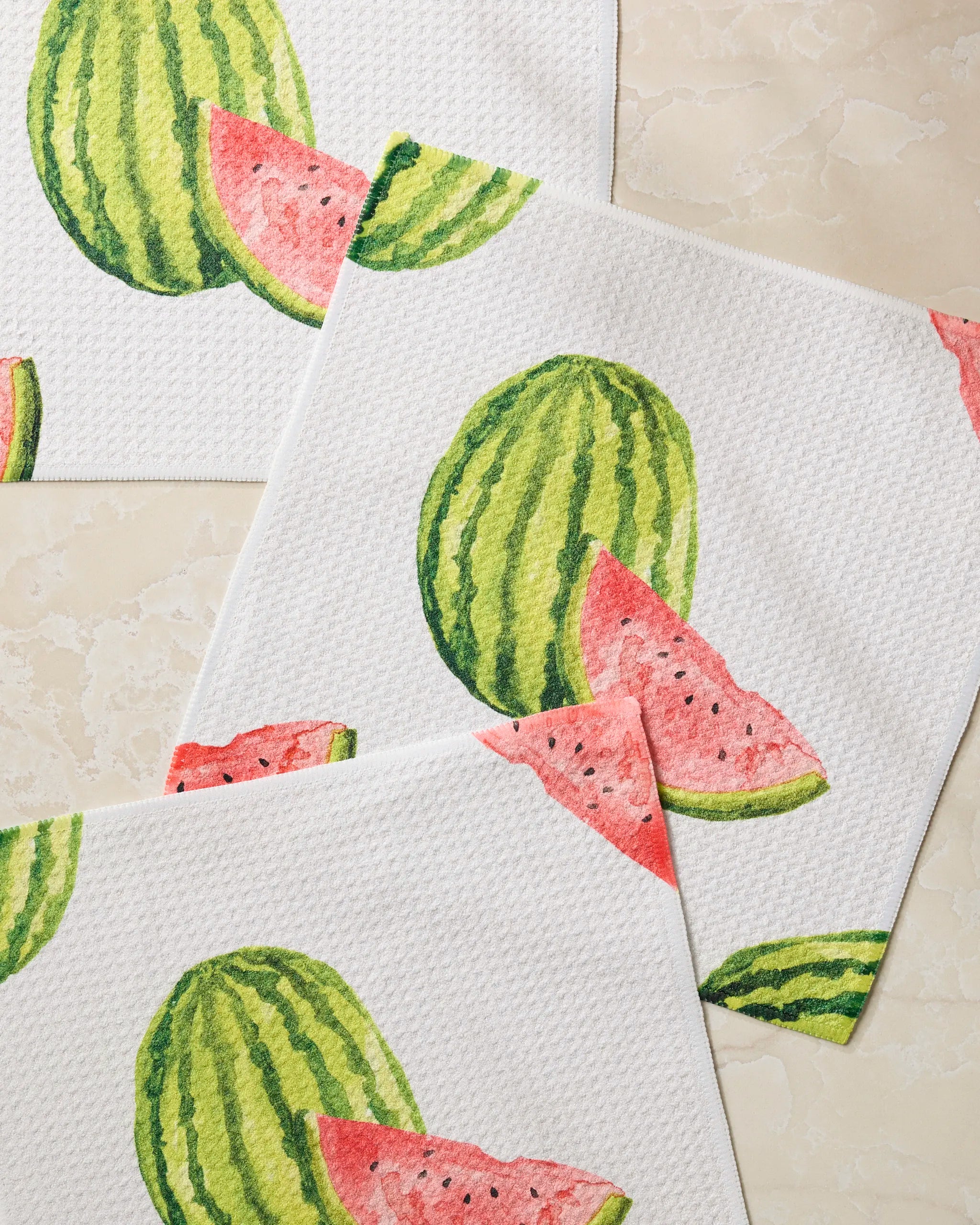 Three “Melon Market” dishcloths by Geometry, made from recycled materials with white textured fabric and watercolor watermelon designs, are overlapping on a light beige marble surface.