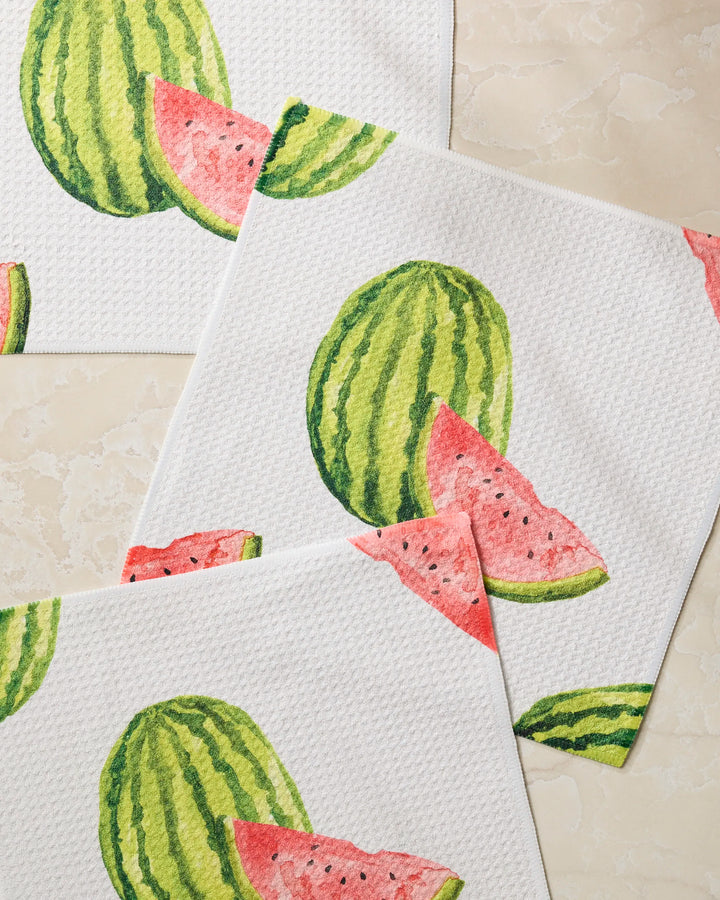 Three “Melon Market” dishcloths by Geometry, made from recycled materials with white textured fabric and watercolor watermelon designs, are overlapping on a light beige marble surface.