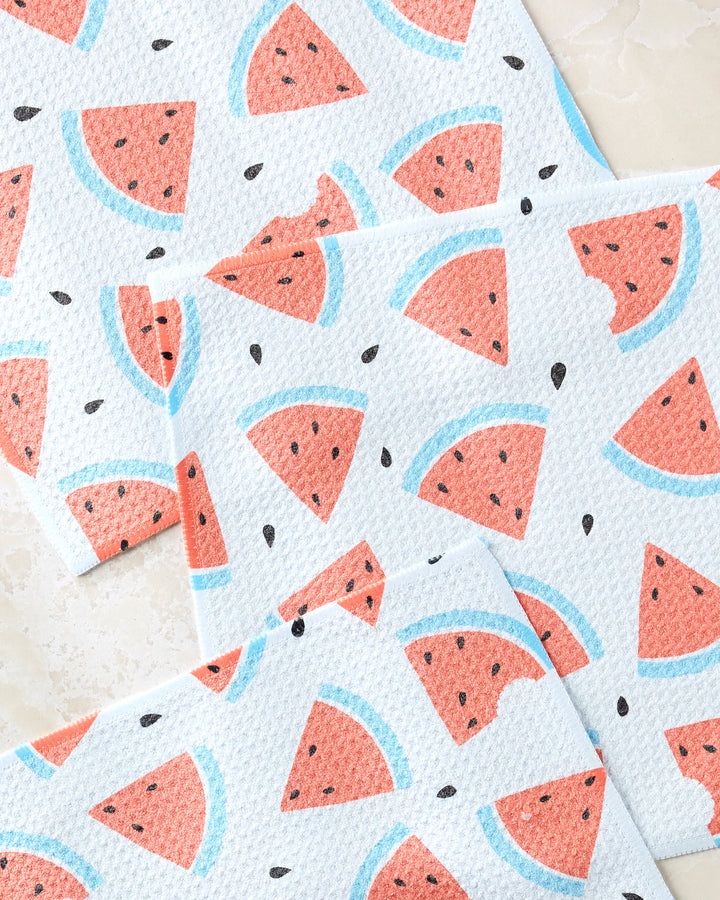 Sara Mari’s Melon Mingle dishtowels showcase a playful watermelon slice pattern—some with bites and scattered seeds—on a white background, overlapping for a cheerful, fresh look.