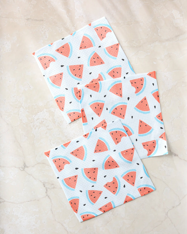 Three white Sara Mari Melon Mingle dishtowels, featuring a repeating watermelon slice pattern, are laid out on a light beige, marble-like surface.