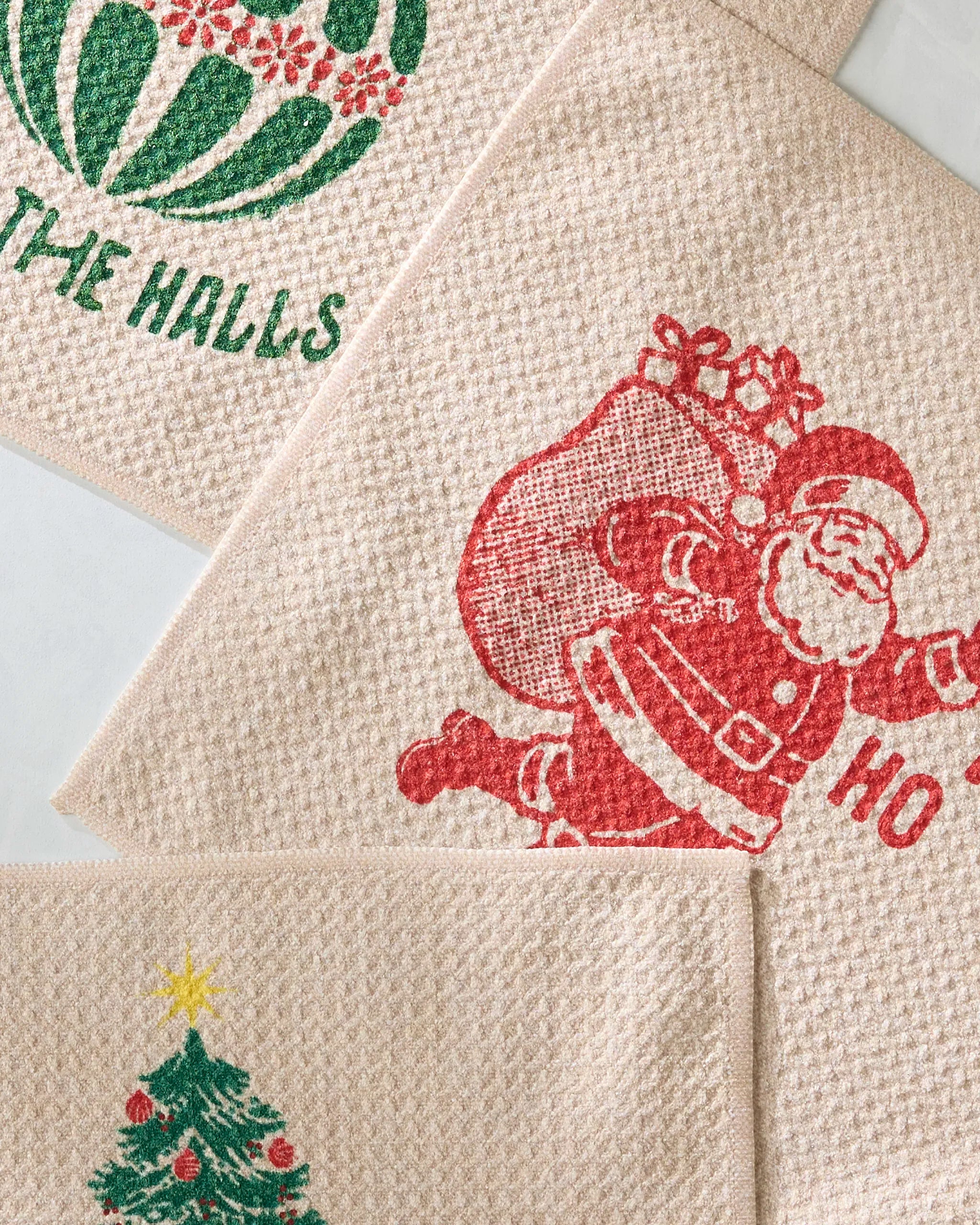 The Geometry Merris set includes three eco-friendly beige dish towels with festive prints: a green ornament with “THE HALLS,” a red Santa with “HO,” and a Christmas tree topped with a yellow star.