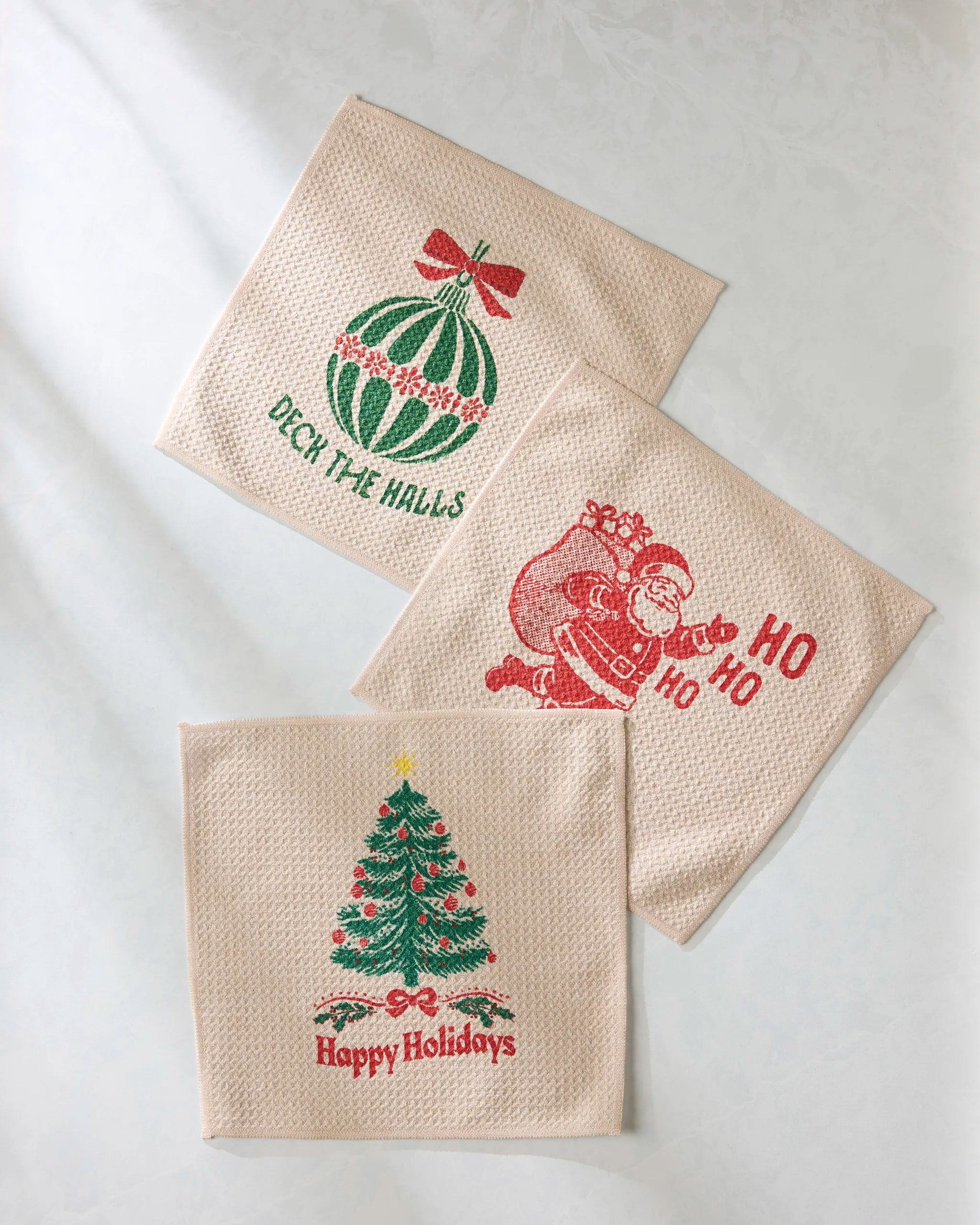 The Geometry Merris set features three beige holiday dishtowels: one with a green ornament and “Deck the Halls,” one showing Santa with “Ho Ho Ho,” and one with a Christmas tree and “Happy Holidays.”.
