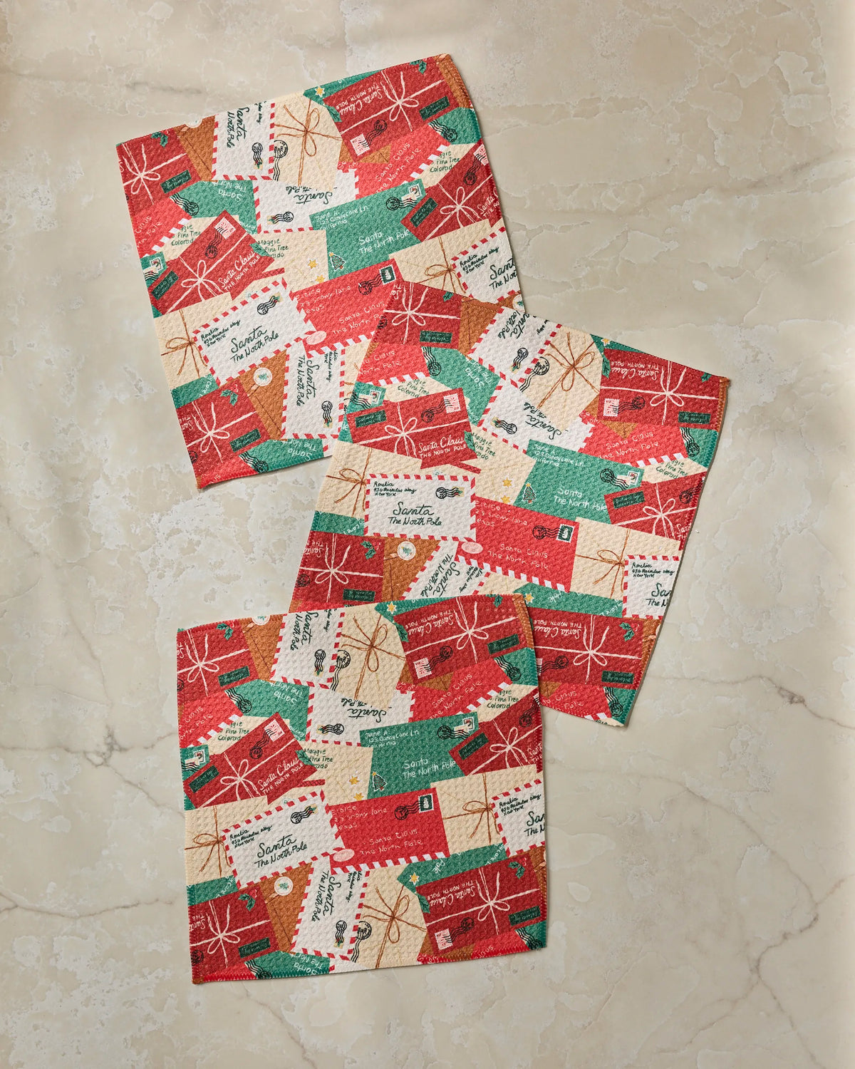 Three square pieces of festive Merry Mail fabric by Geometry, featuring red, green, and beige holiday-themed designs with gift tags and bows, are arranged on a light marble surface. This eco-friendly dishcloth set is crafted from recycled materials to add a sustainable touch to your kitchen décor.