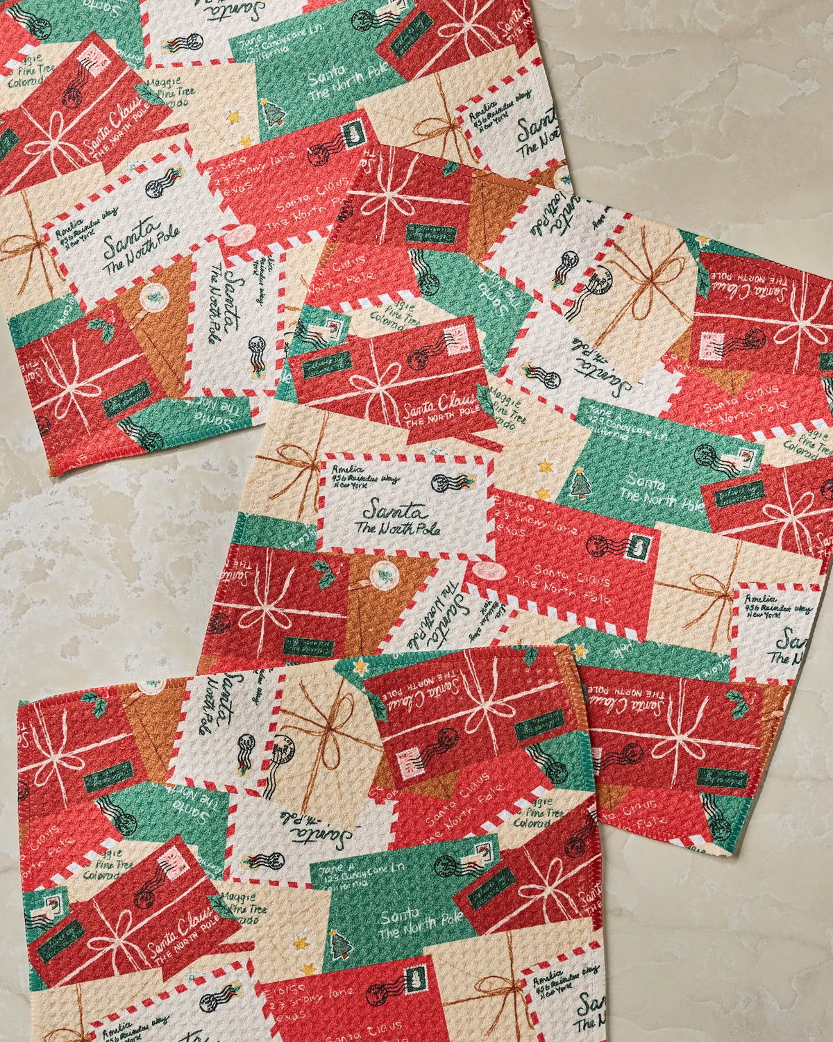 Three “Merry Mail” paper napkins from Geometry, made from recycled materials and featuring a festive pattern of red, green, and white envelopes and parcels with holiday-themed text like Santa, The North Pole, and decorative postage stamps, are arranged on a light-colored surface.