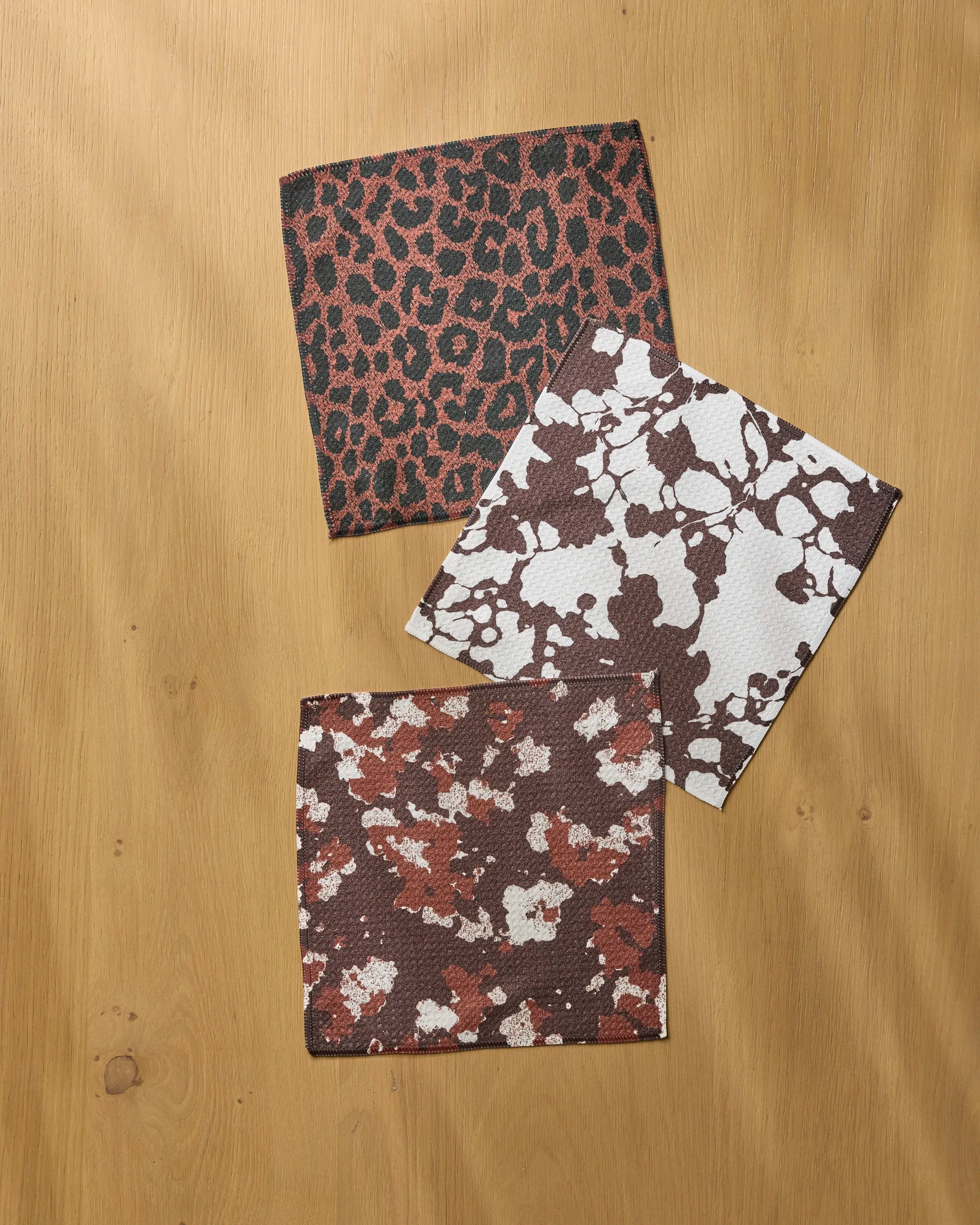 Three square fabric swatches with animal prints—crafted from recycled materials—are displayed on light wood, showcasing the bold, eco-friendly style of Geometry's Midnight Vine Noir dishcloth set.