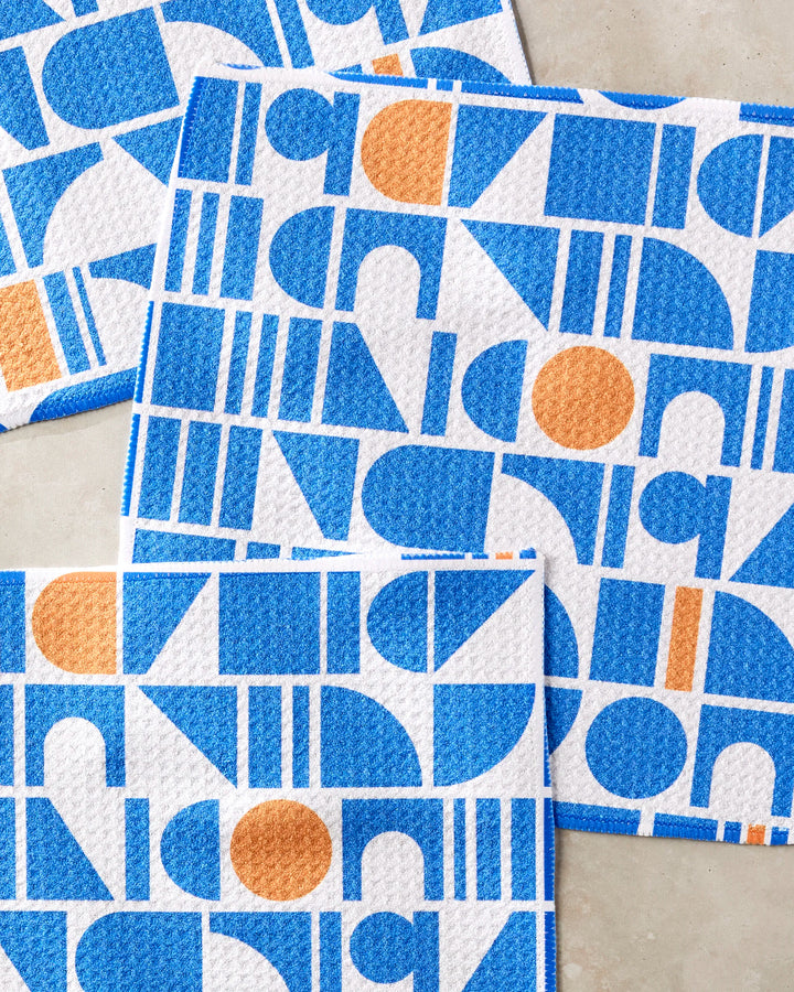 Four Misty Oasis dishtowels by Happy Hemera, featuring bold blue and orange geometric patterns—circles, semi-circles, and rectangles—are laid out on a beige surface, overlapping at the corners.