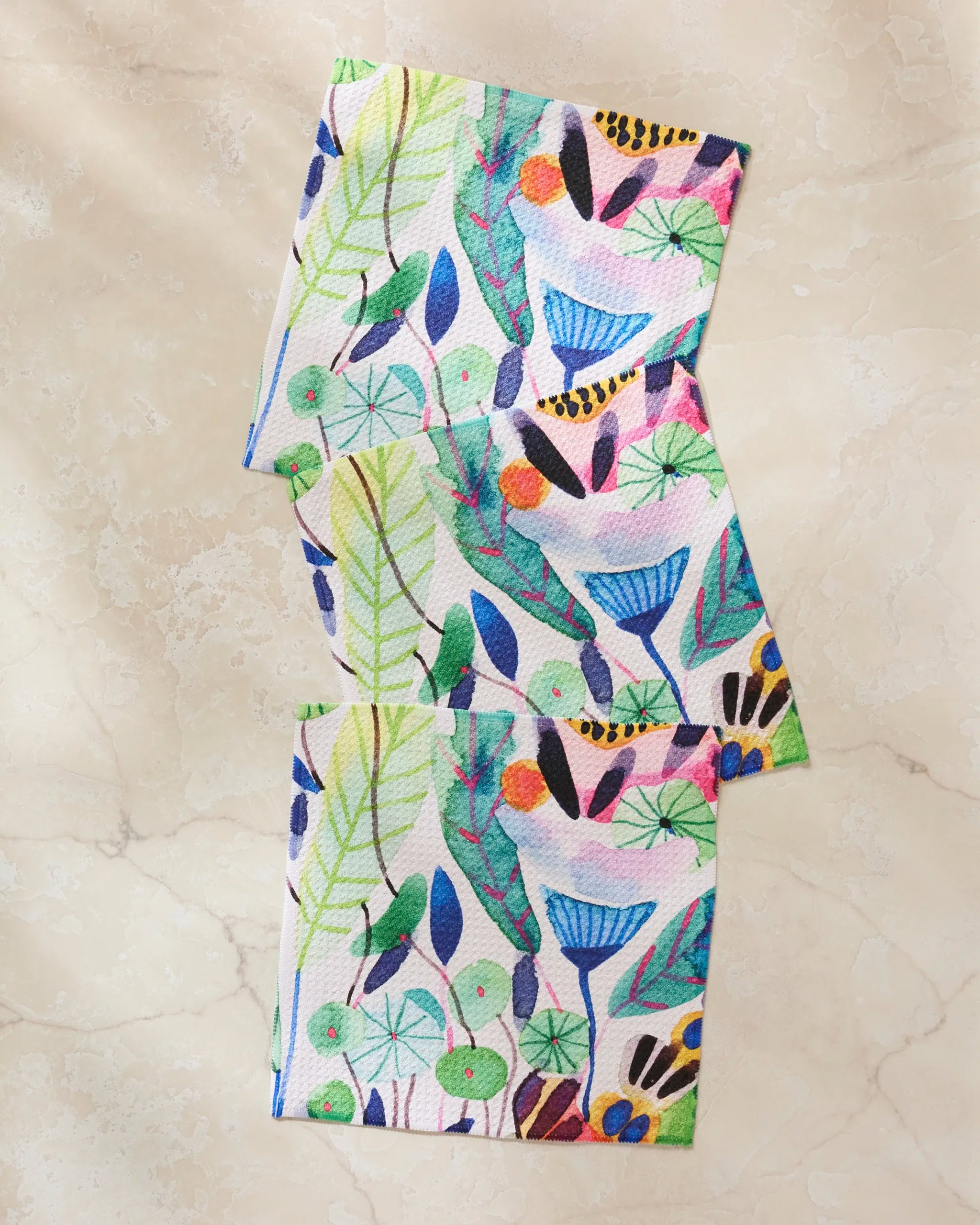 Three "Morning" napkins by Maria Montiel, made from recycled materials, feature a lively nature-inspired design with leaves, flowers, berries, and birds displayed on a light marble surface.