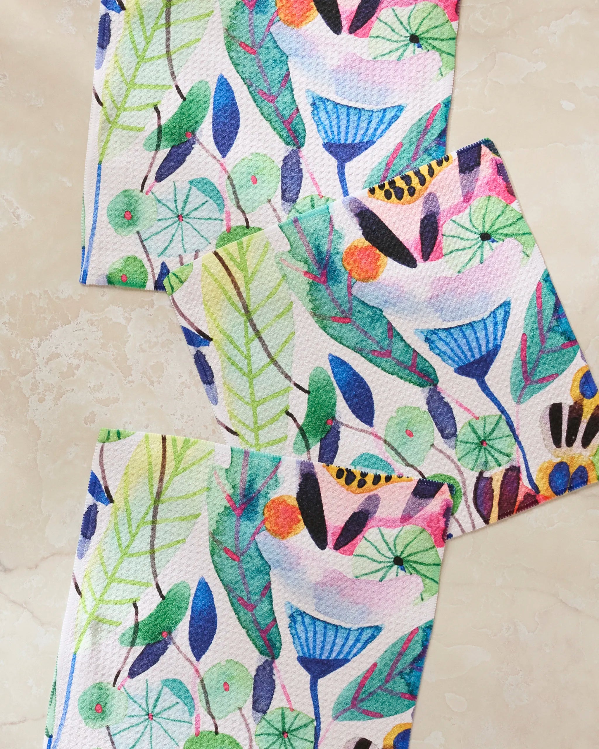 Three "Morning" paper napkins by Maria Montiel, featuring colorful abstract botanical designs, are arranged diagonally on a light beige marble surface—an eco-friendly choice made from recycled materials.