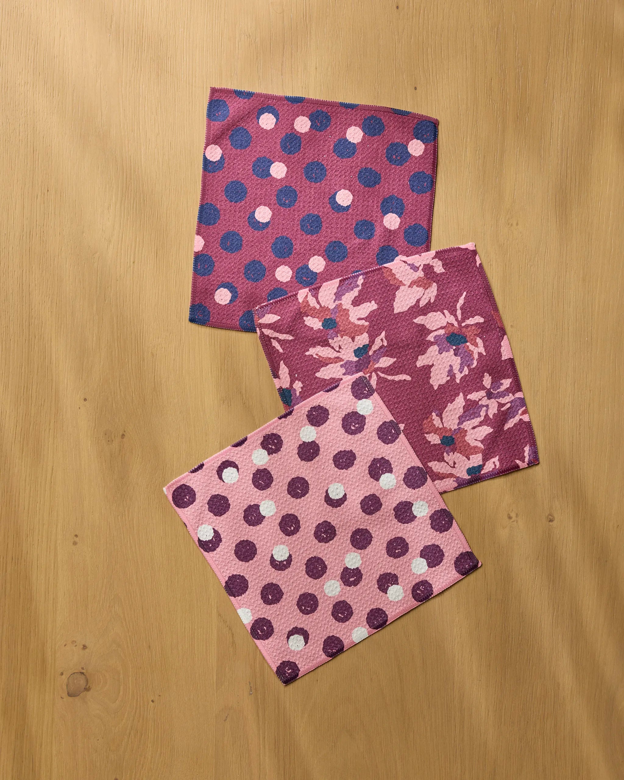 The Geometry Mulberry Bloom Dot set includes three square napkins—two with purple and pink polka dots and one with a floral design—arranged on light wood for a stylish, functional addition to any kitchen.