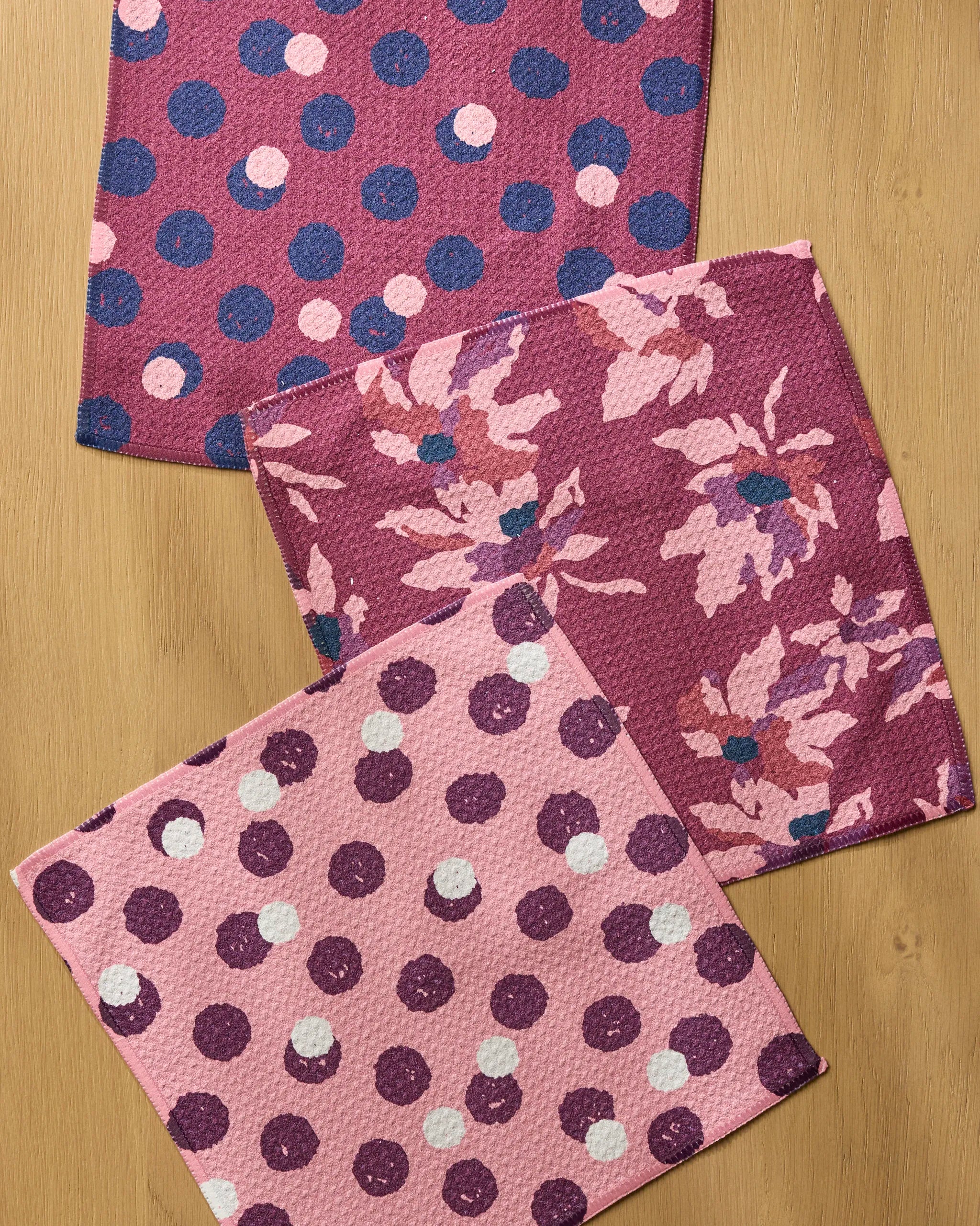 Three Geometry Mulberry Bloom Dot dishcloths rest on a wooden surface—two with playful polka dots, the third with a pink and magenta floral pattern—from the colorful recycled set.